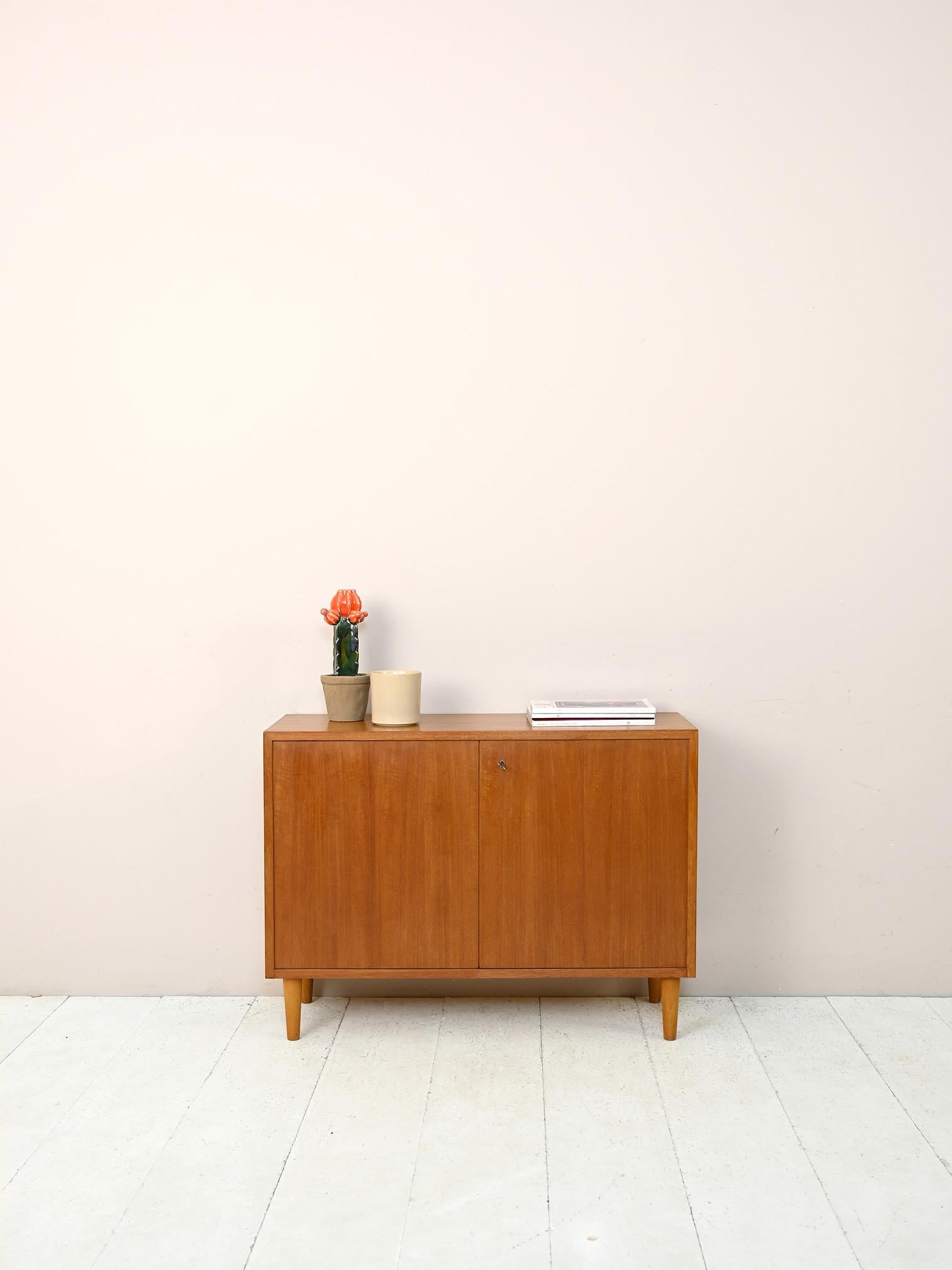 Small Scandinavian sideboard from the 1960s.

Simple forms reminiscent of mid-century modern taste.
The structure with square lines consists of a storage compartment closed by lockable hinged doors. Tapered, light oak wood feet lighten the