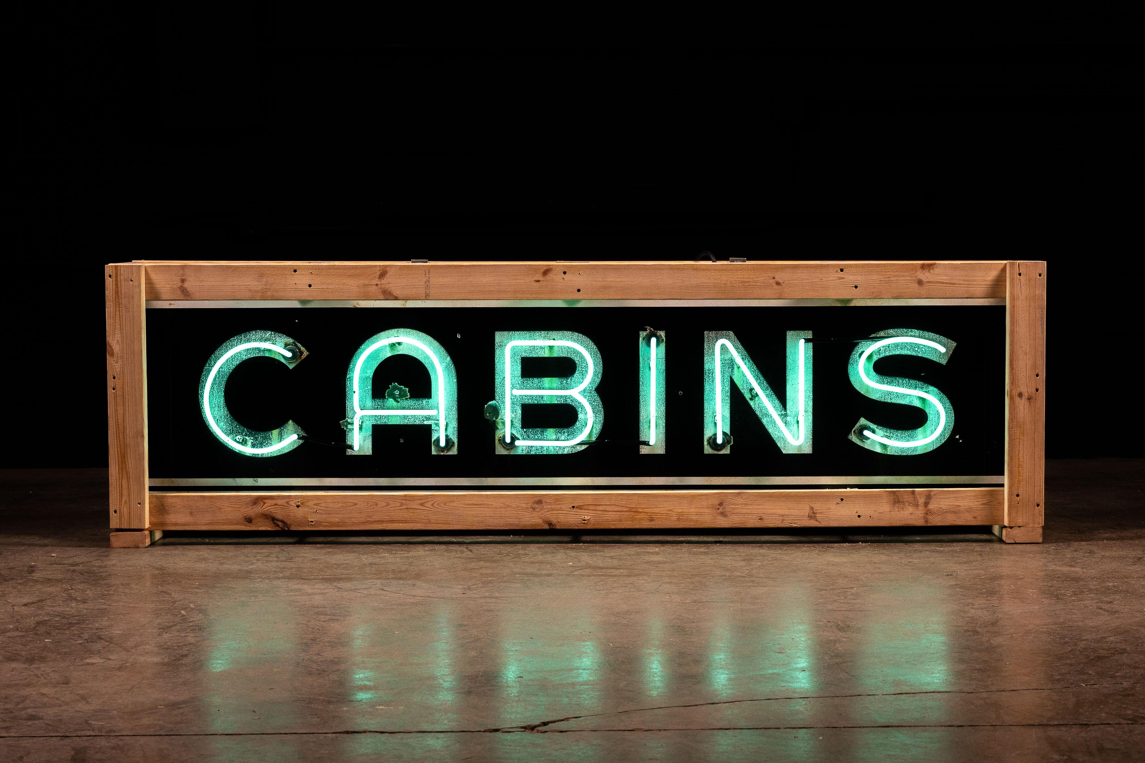Vintage neon sign advertising cabins for rent. This piece features a bold Bauhaus-style font in white set against a dark background. The lettering has been traced in a turquoise neon which gives high contrast against the porcelain's dark