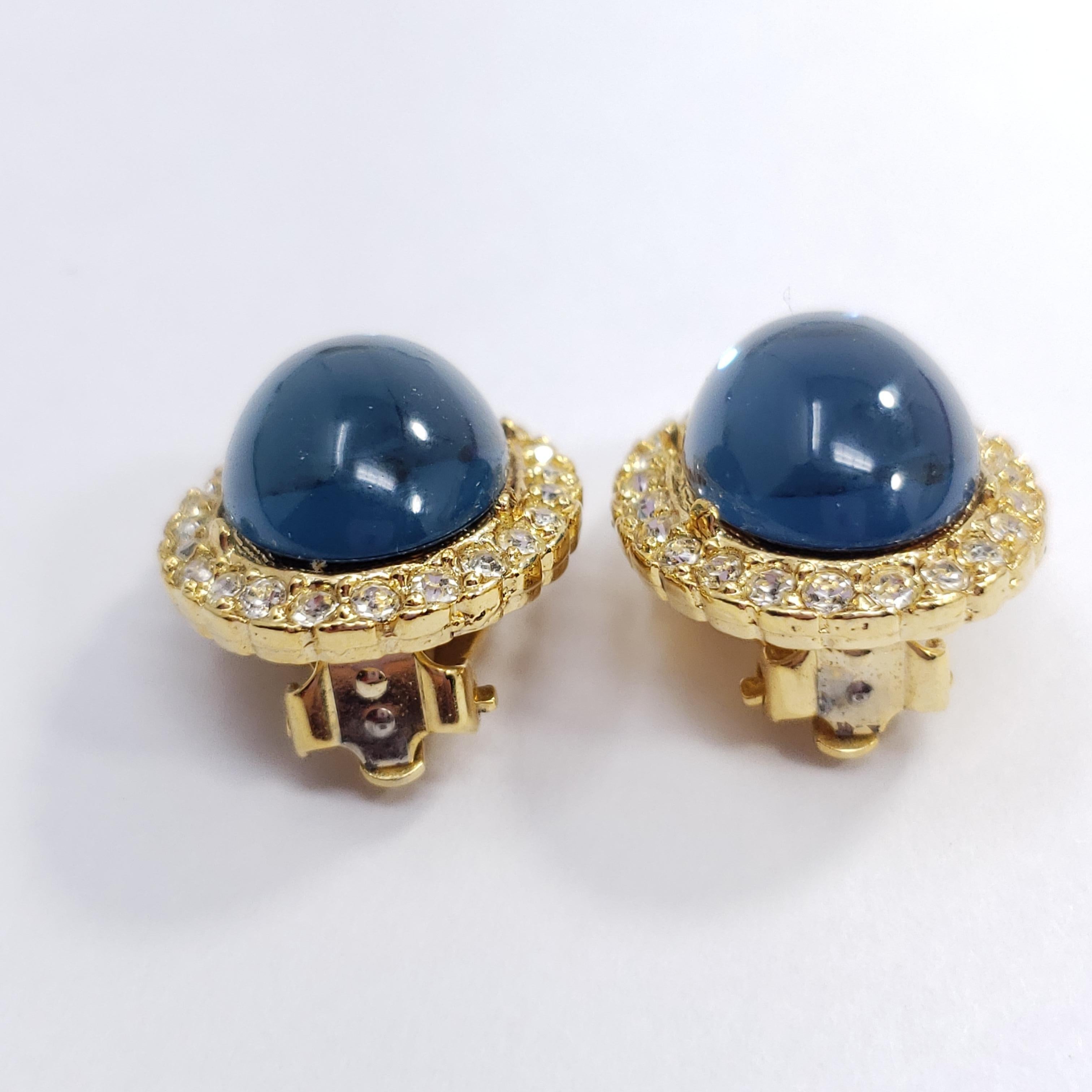 A pair of  exquisite clip on earrings, each featuring a single, royal, sapphire-blue cabochon accented with smaller crystals, set on a golden clip.

Gold-filled. Made in the USA. Circa mid to late 20th century.

Each earring approx. 1 inch by 0.7