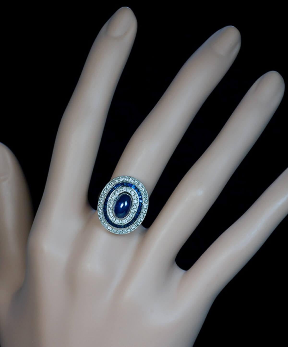 Belgium, 1940s  The ring is handcrafted in 18K white gold. It is centered with a cabochon cut blue sapphire encircled by two rows of diamonds (about 1 carat total weight) and a row of channel-set French cut sapphires.  The cabochon sapphire measures