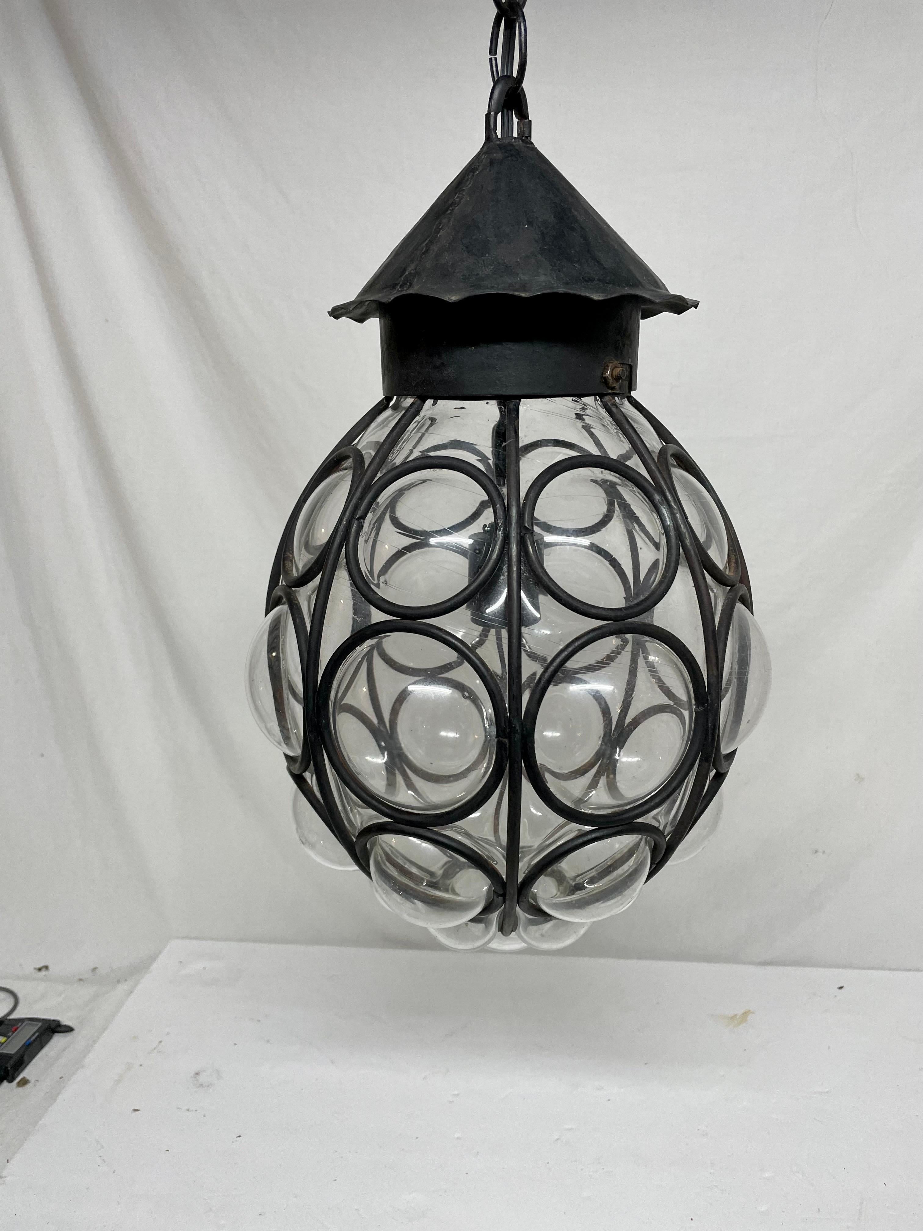 Caged glass lantern with single medium-base (Edison) light. Original black painted wrought iron with transparent glass piece encased by an iron honeycomb style frame. In this style of light the glass is generally hand blown inside the cage to create