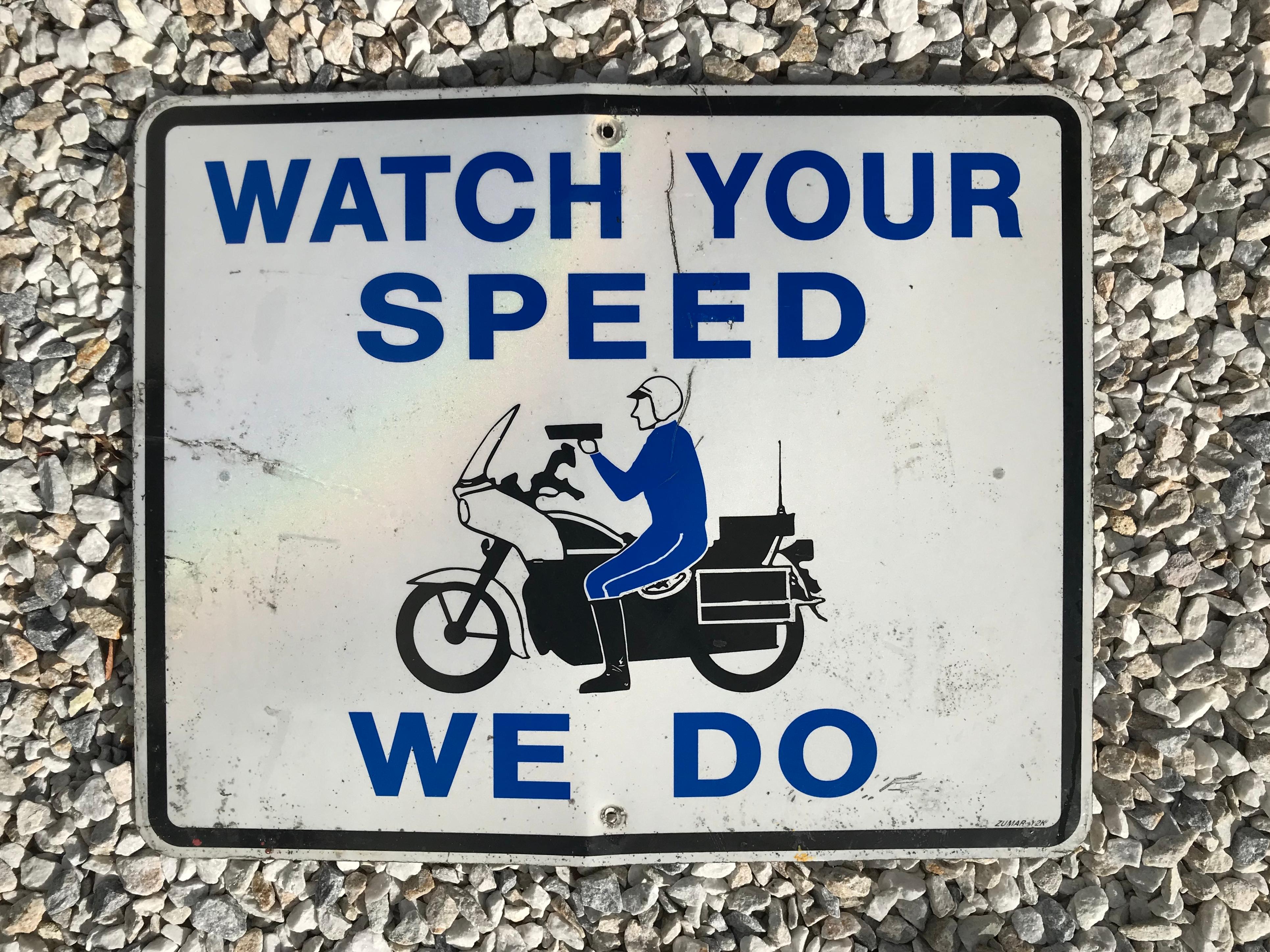 Cool vintage California highway sign. Sign reads - Watch Your Speed We Do featuring a police officer riding a motorcycle in the middle. White reflective steel sign with blue lettering. Great graphics. Some bends to metal and staining on front.