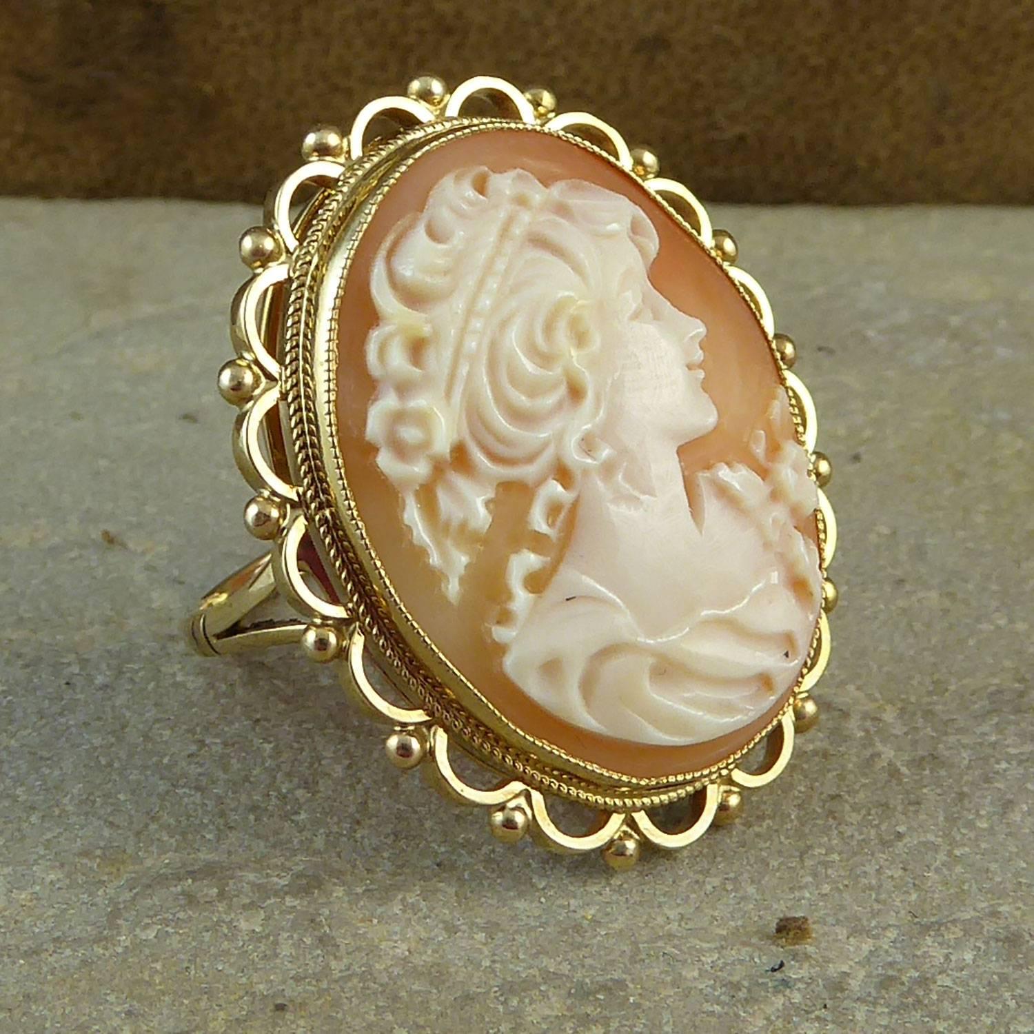 A pretty vintage cameo mounted in a ring setting and creating a ring of quite impressive size.   The cameo has been carved to show a pretty young woman with a a flowing up-style hair-do and a flower pinned to her dress.  The setting in yellow gold