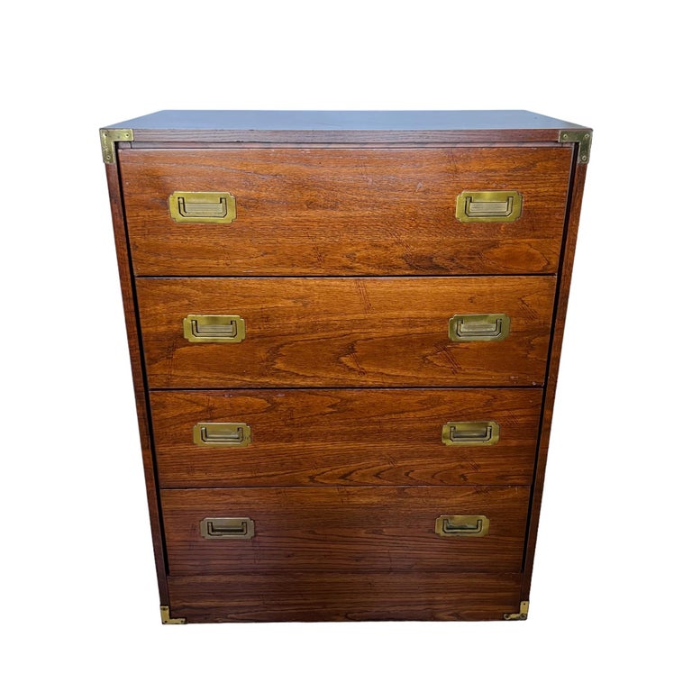 A Mid-Century Modern Campaign style four drawer highboy dresser circa 1990. Relatively small in size, it features solid wood framing with high quality walnut veneer & durable wood laminate top surface, solid brass handles & plated metal corner