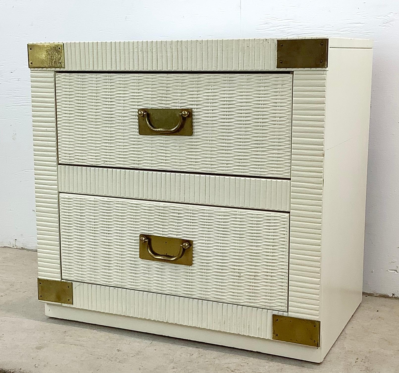 Invite the glamour of a bygone era into your bedroom with this vintage Hollywood Regency two-drawer nightstand. Its crisp white wicker is intertwined with the luxurious sheen of brass campaign hardware, encapsulating the extravagance of it's chic