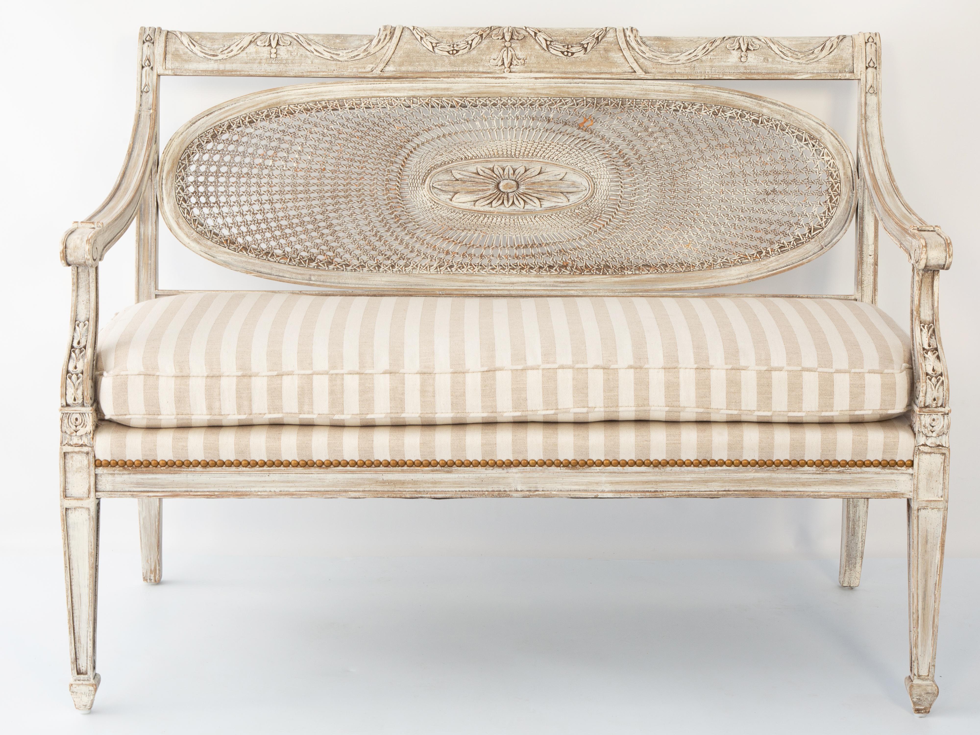 Settee, having a painted frame showing natural wear. Its crestrail carved with swags, over oval caned backrest, centered with an oval rosette. Downswept arms ending in scrolls, on foliate-carved terminals. Its loose cushion seat on upholstered deck,