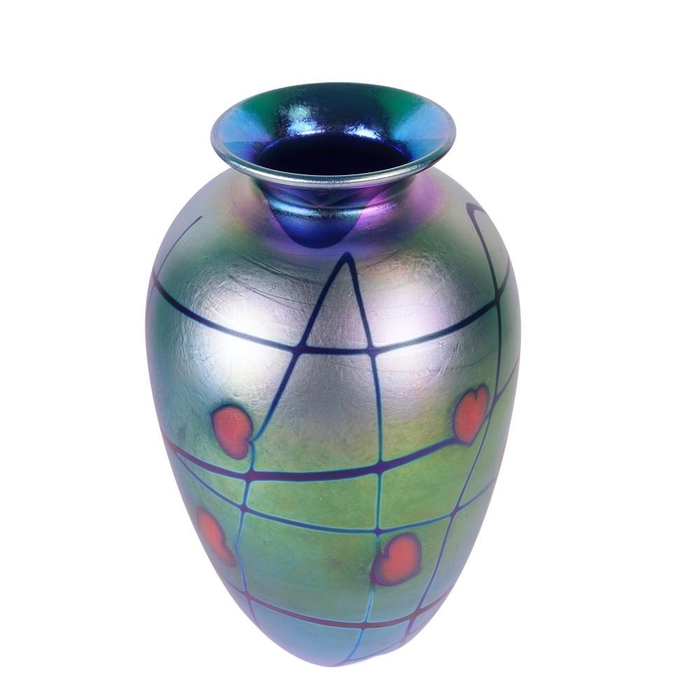 Presenting this outstanding Donald Carlson art glass vase. Vase is decorated with red hearts and dark blue vines, known as a “heart and vine” design. The main color of this vase is blue with peacock iridescence displaying purple, blue, silver and