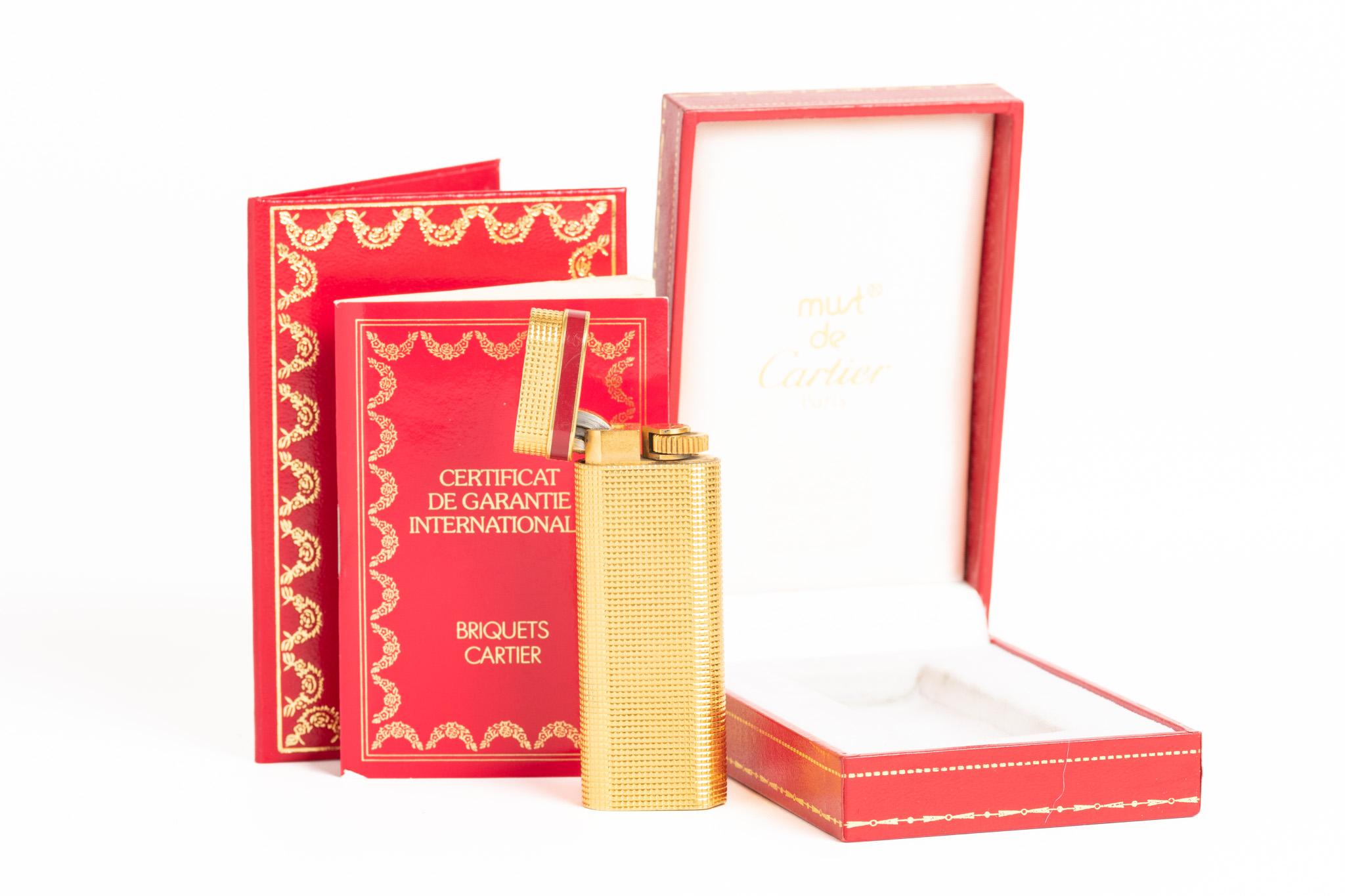 A rare and classic gold-plated Cartier lighter with a red enameled band at the top. Inscribed with 'Cartier Paris' along the bottom edge with a serial number: 94263V. The lighter has a beautiful geometric pattern. The lighter comes with an original