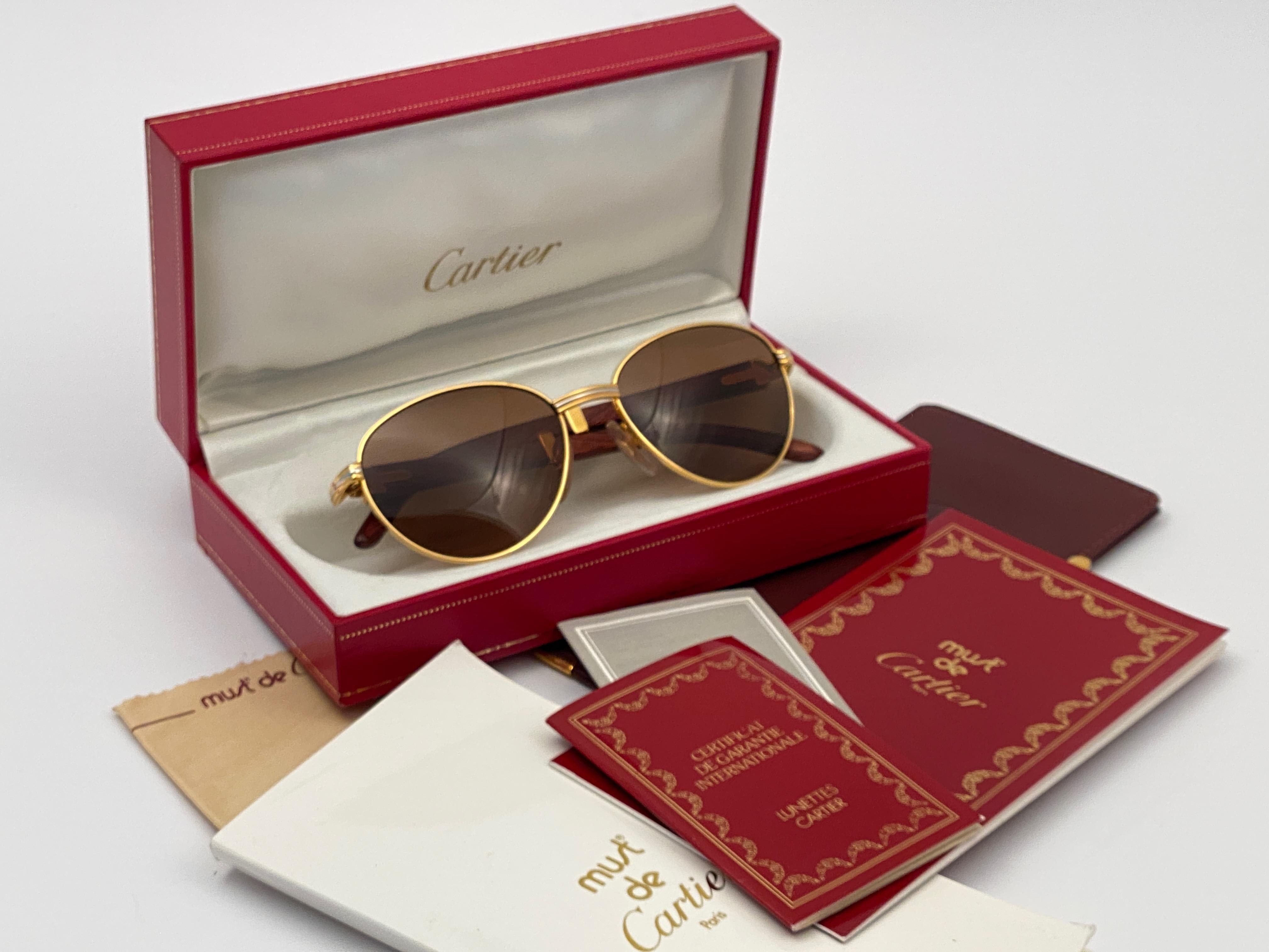 Nuevas gafas de sol Cartier 1990 Full Set Auteuil Hardwood con lentes nuevas de color marrón miel sólido (protección uv). 
El marco es con el anverso y los laterales en oro amarillo y blanco y tiene las famosas patillas con acentos de madera y oro.