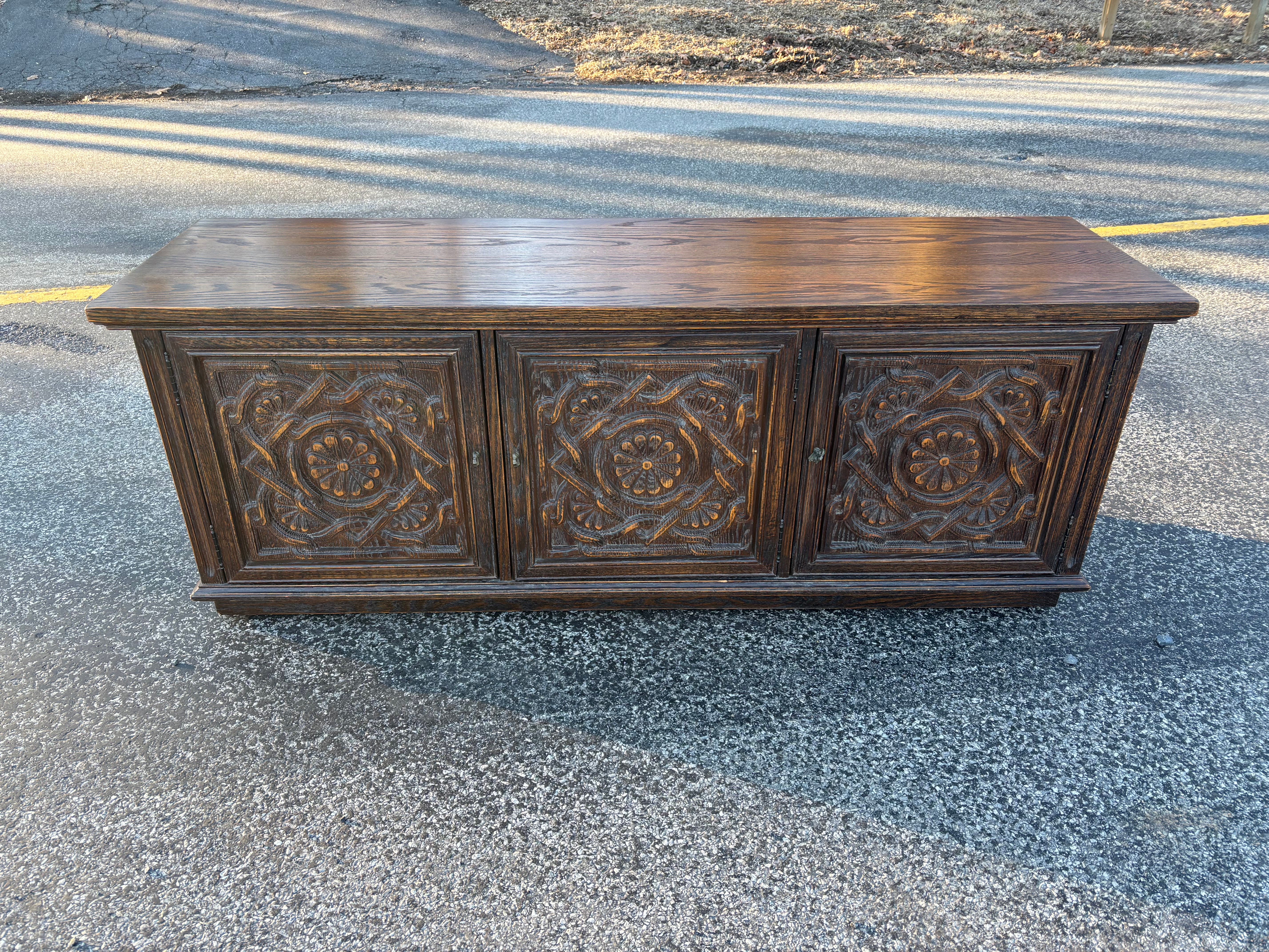 Buffet/crédence en chêne massif féodal, armoire à 3 portes.  Comprend deux portes à gauche qui s'ouvrent sur une étagère et une porte à droite qui comporte deux tiroirs coulissants et un espace ouvert en dessous.  Magnifique motif floral sculpté sur