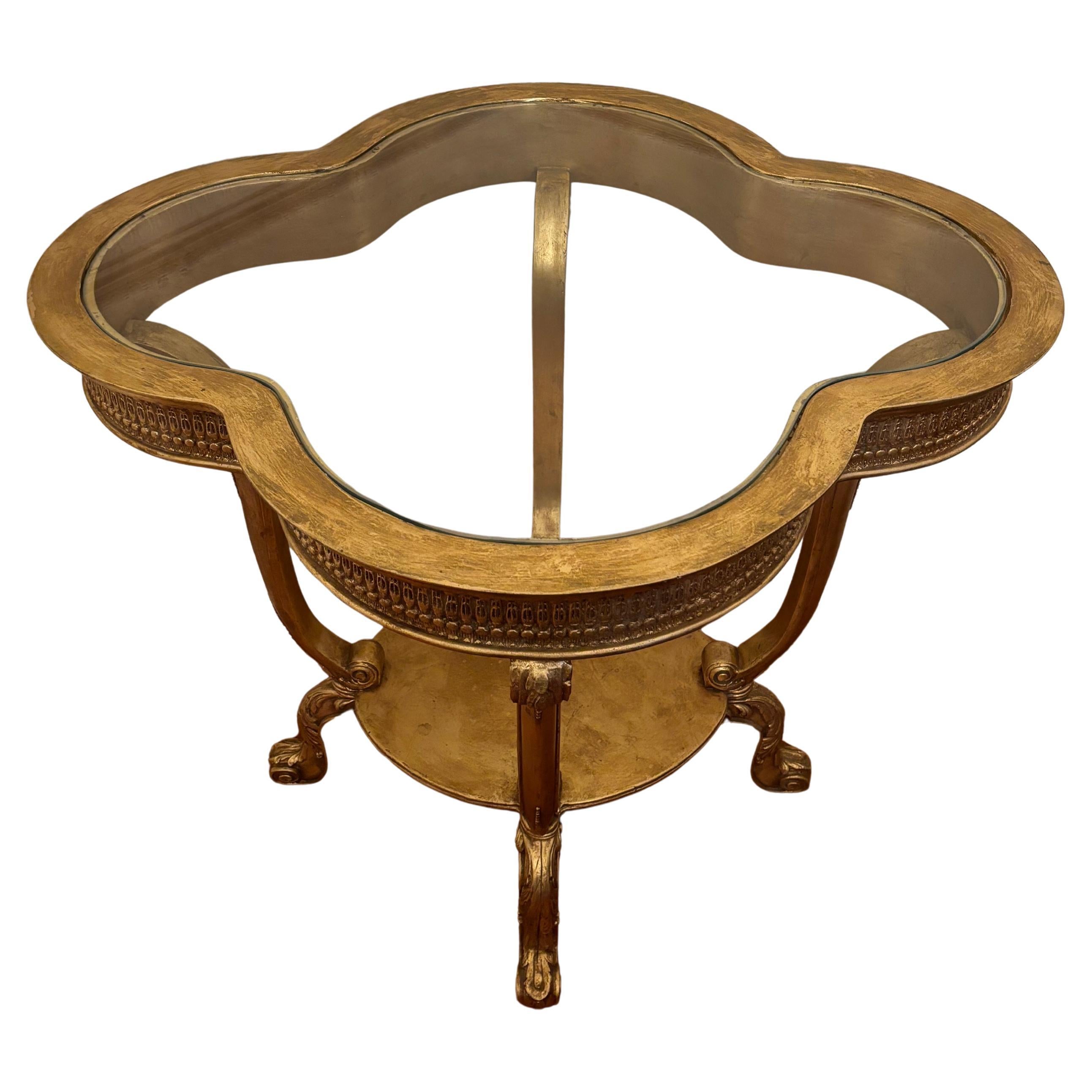 Il tavolo Wood Brothers Hermitage è in stile Luigi XV e presenta una forma a quadrifoglio in legno dorato intagliato con vetro incastonato. Il grembiule delicatamente intagliato poggia su quattro gambe cabriole collegate da una base centrale. La