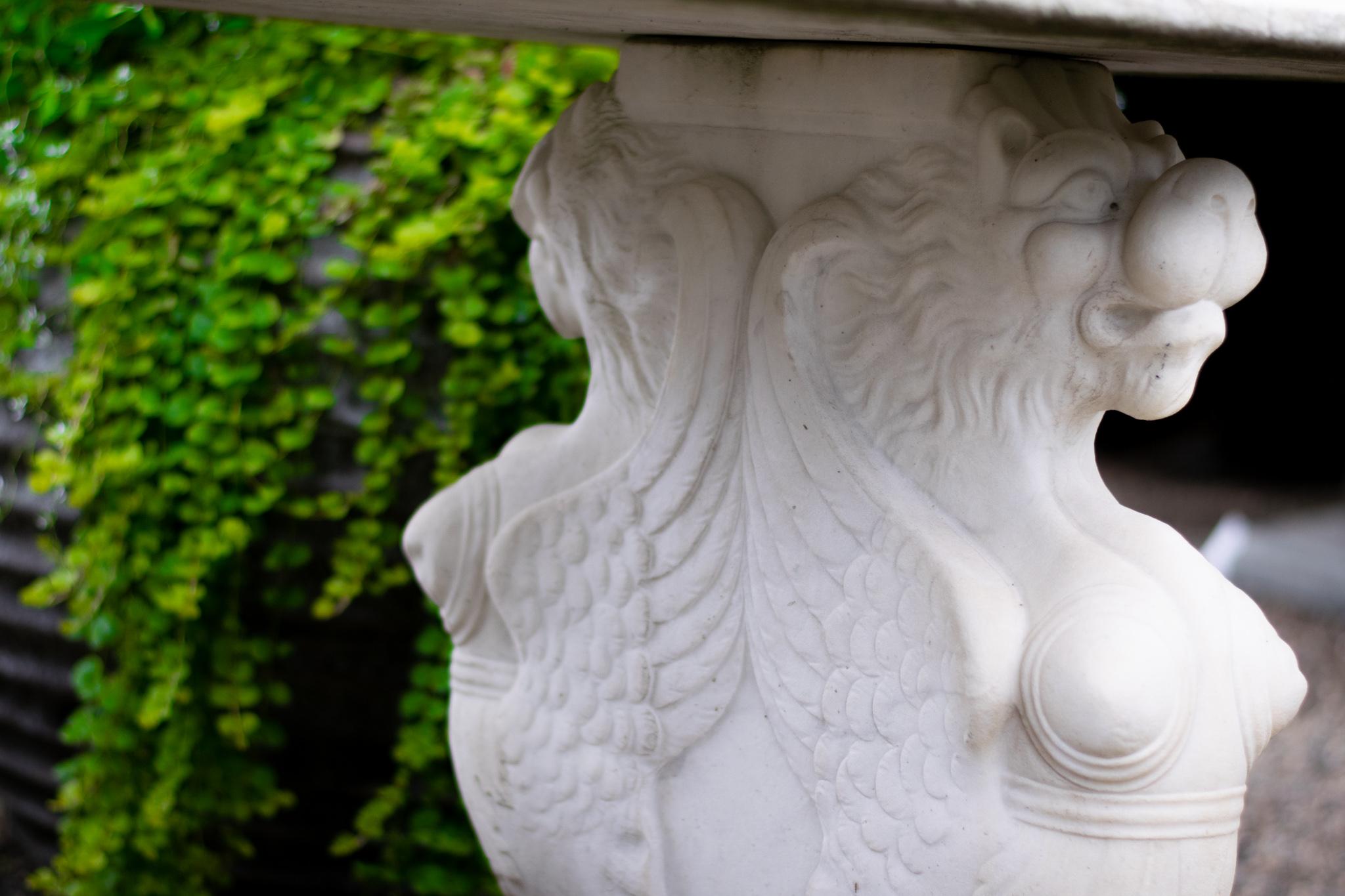 A wonderfully carved vintage marble table featuring an intriguing botanical pattern on the top surrounding an anonymous face, supported by an impressive pair of winged lions feet. Poised to be the center of attention, this stunning white table will