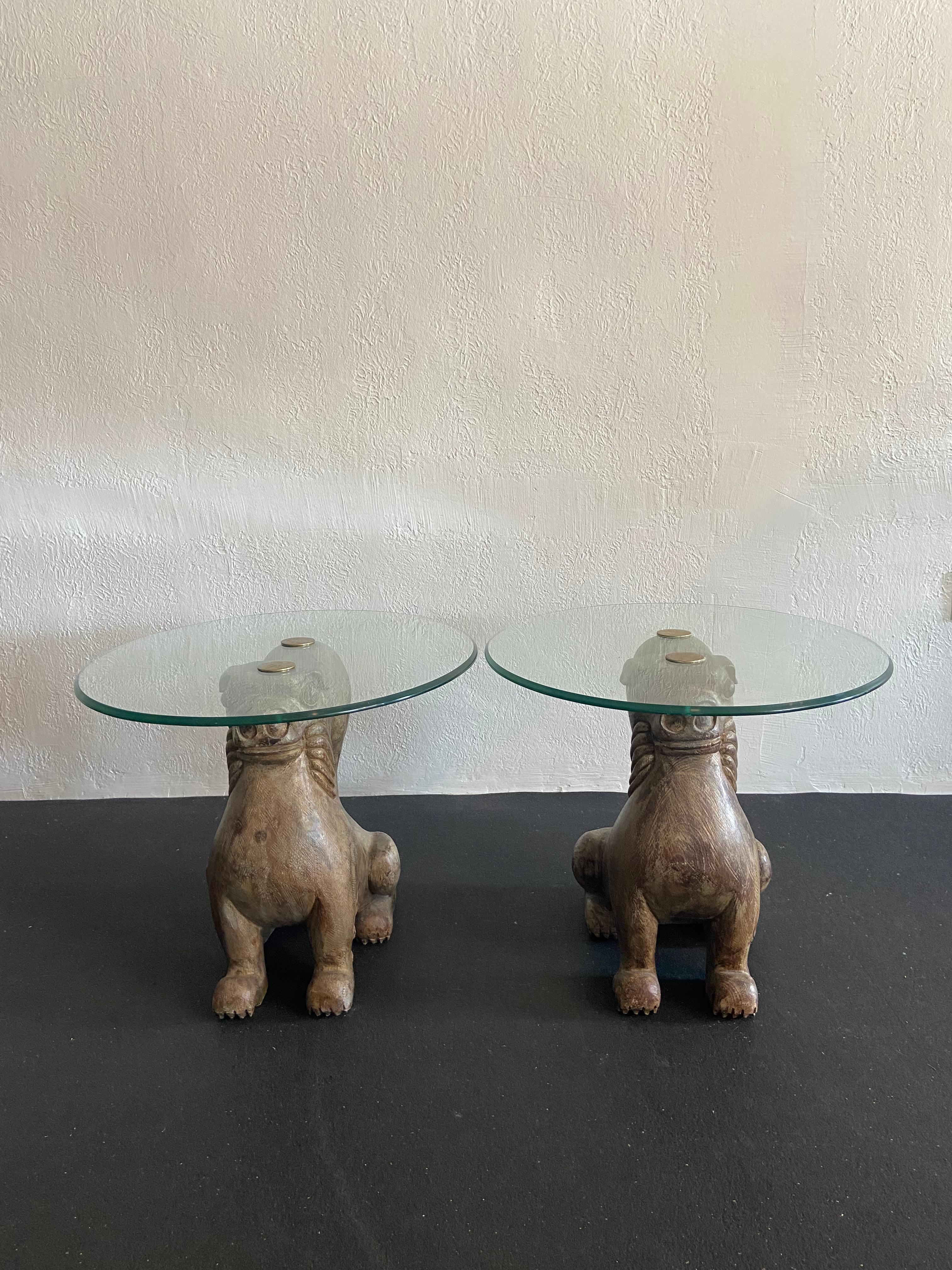 Pair of carved wood and brass foo dog end tables by Sarreid Ltd. Patina to the brass. Wear to the as found vintage glass tops. 

Would work well in a variety of interiors such as modern, mid century modern, Hollywood regency, etc. Piece blends