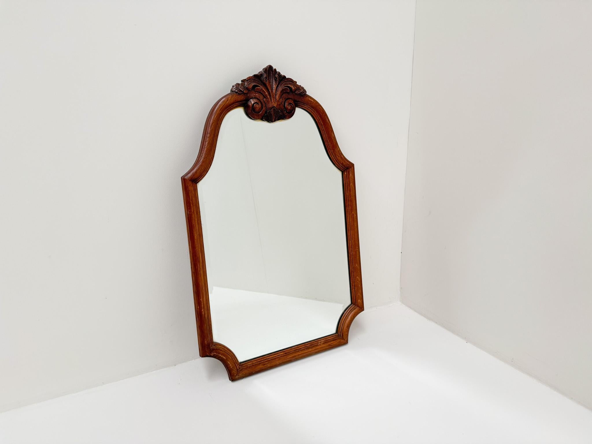 Elegant miroir mural vintage doté d'un cadre en bois massif avec de magnifiques acanthes sculptées à la main au niveau de la crête. Le cadre met en valeur des tons naturels chauds et un travail artisanal fin, tandis que le verre biseauté ajoute une