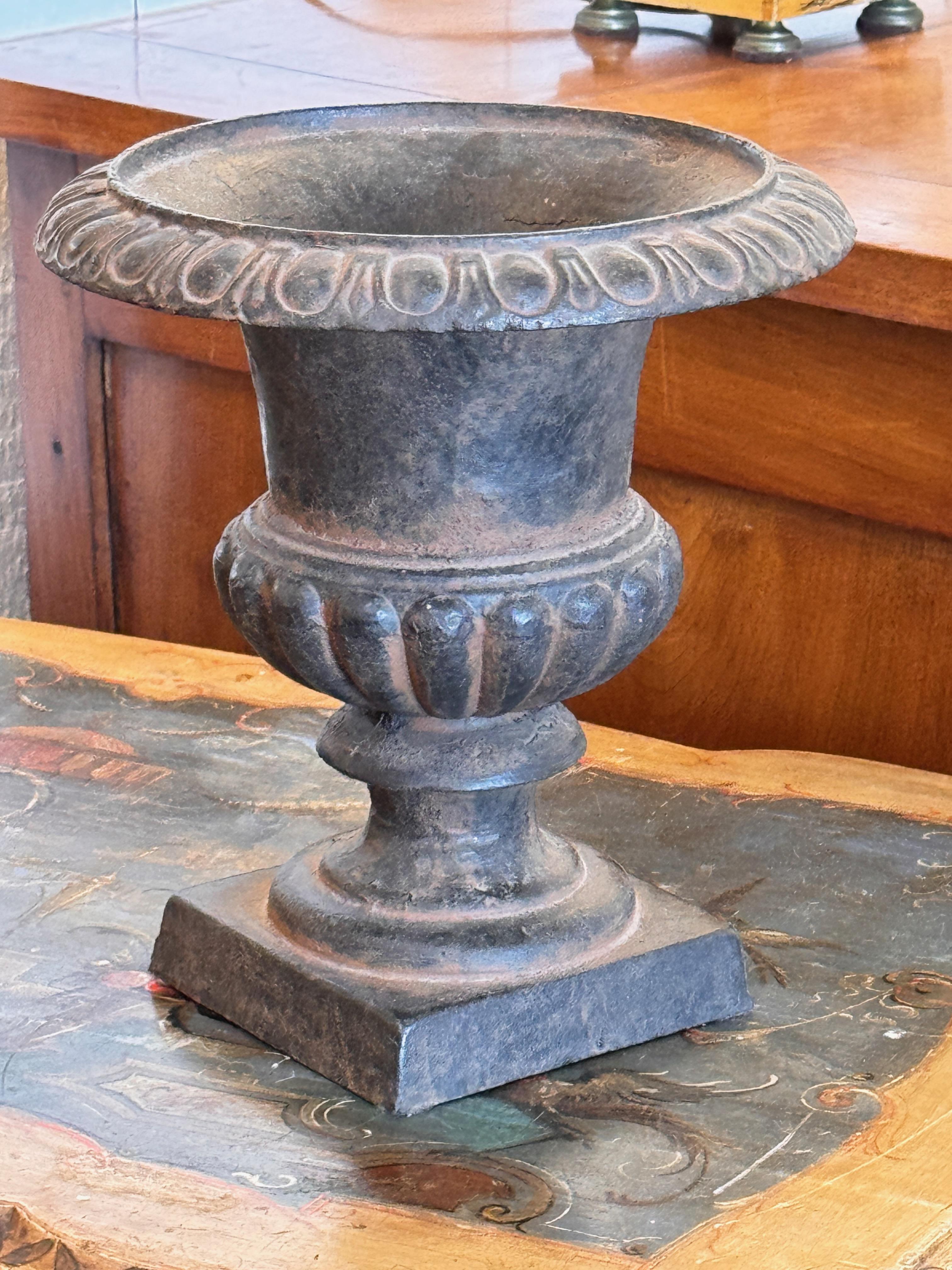 Urne en fonte lourde avec un large bord évasé, une partie centrale lobée et une base carrée. La surface vieillie présente une altération subtile, caractéristique de la ferronnerie d'époque. Forme classique avec cannelures décoratives autour du col