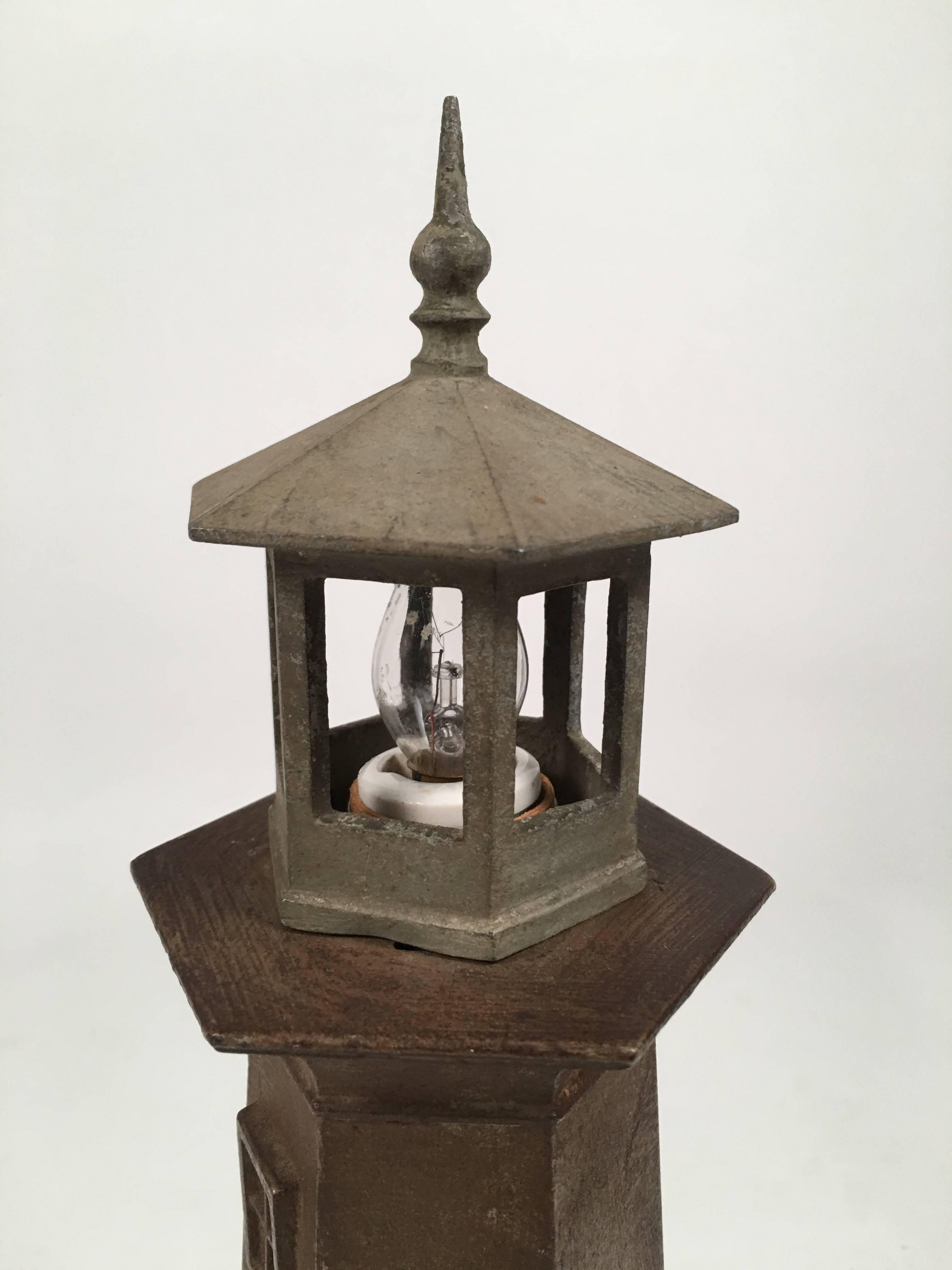A vintage, well modeled, cast metal lighthouse lamp, the hexagonal lantern with finial on top housing a ceramic socket with a night light bulb, over the hexagonal tower with windows on various levels, with a central door just above the stepped, wood