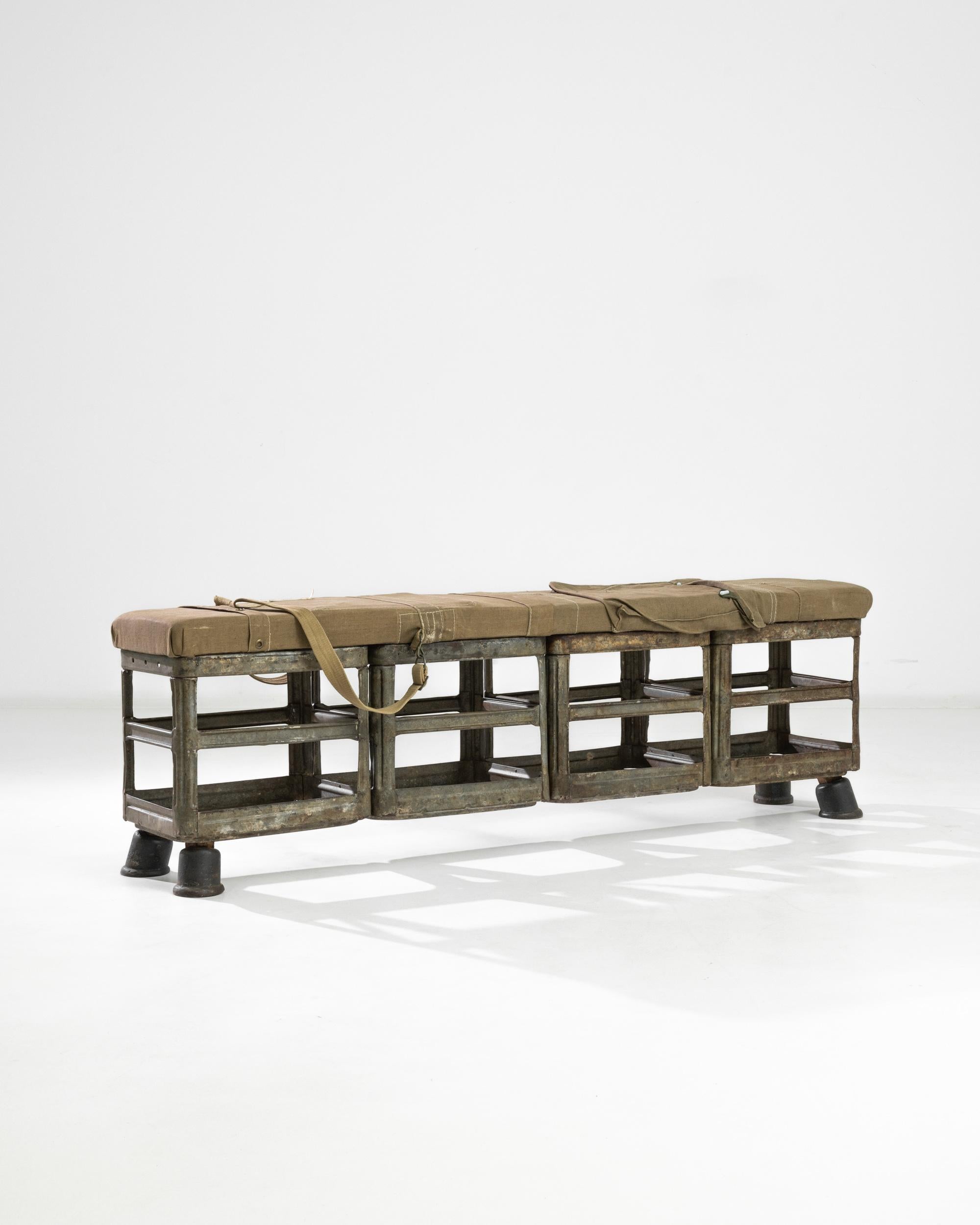 A metal bench with upholstered seat from Central Europe, produced during the 20th century. Four metal crates stacked sideways and resting on four lightly angled feet, topped by an olive-beige upholstered cushion. Looking like vintage military