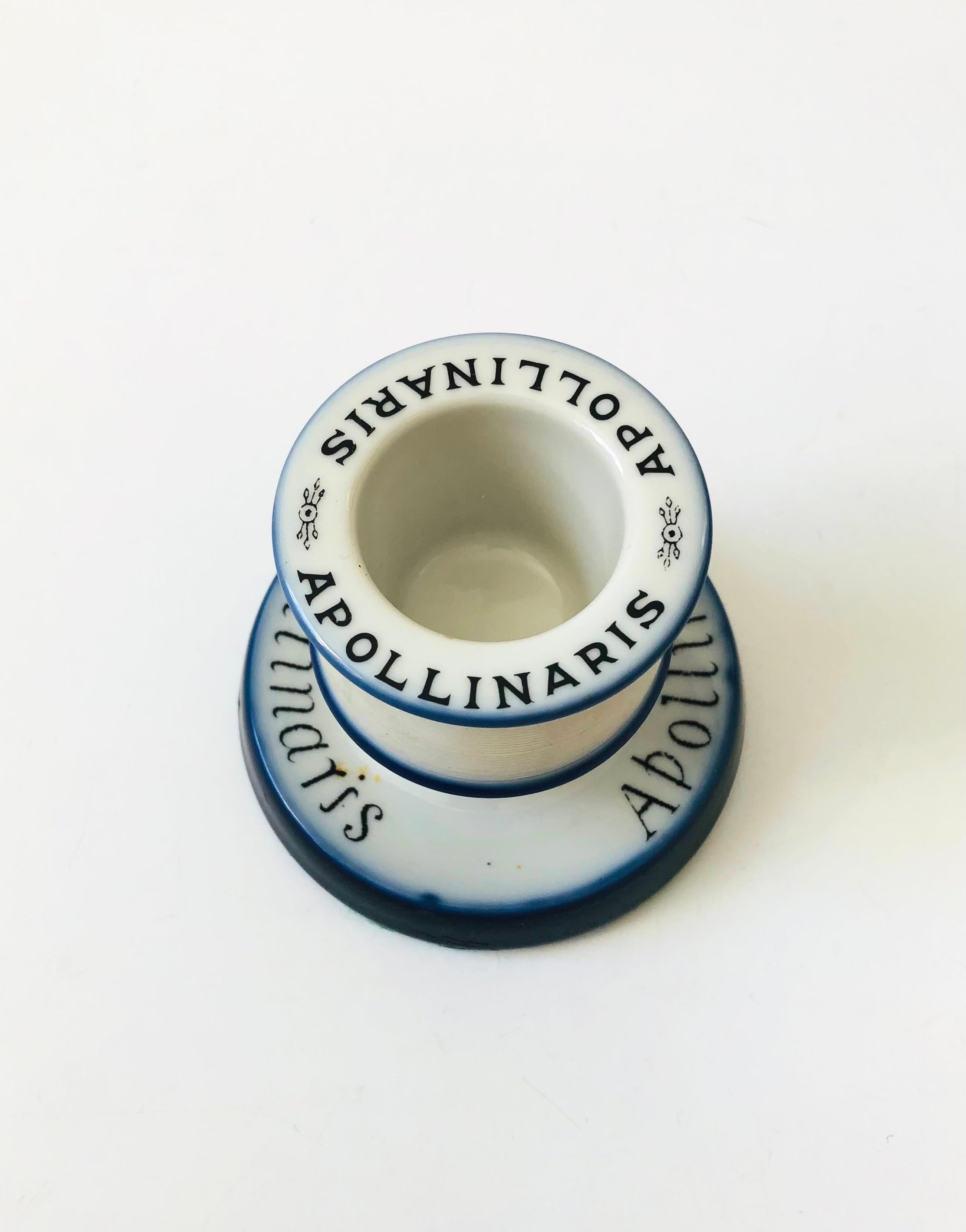 A vintage ceramic match striker and holder advertising Apollinaris Mineral Water. Features a compartment for holding matches with a ribbed texture to the ceramic for using as a striker. Glossy white glaze with cobalt blue banding. Produced in