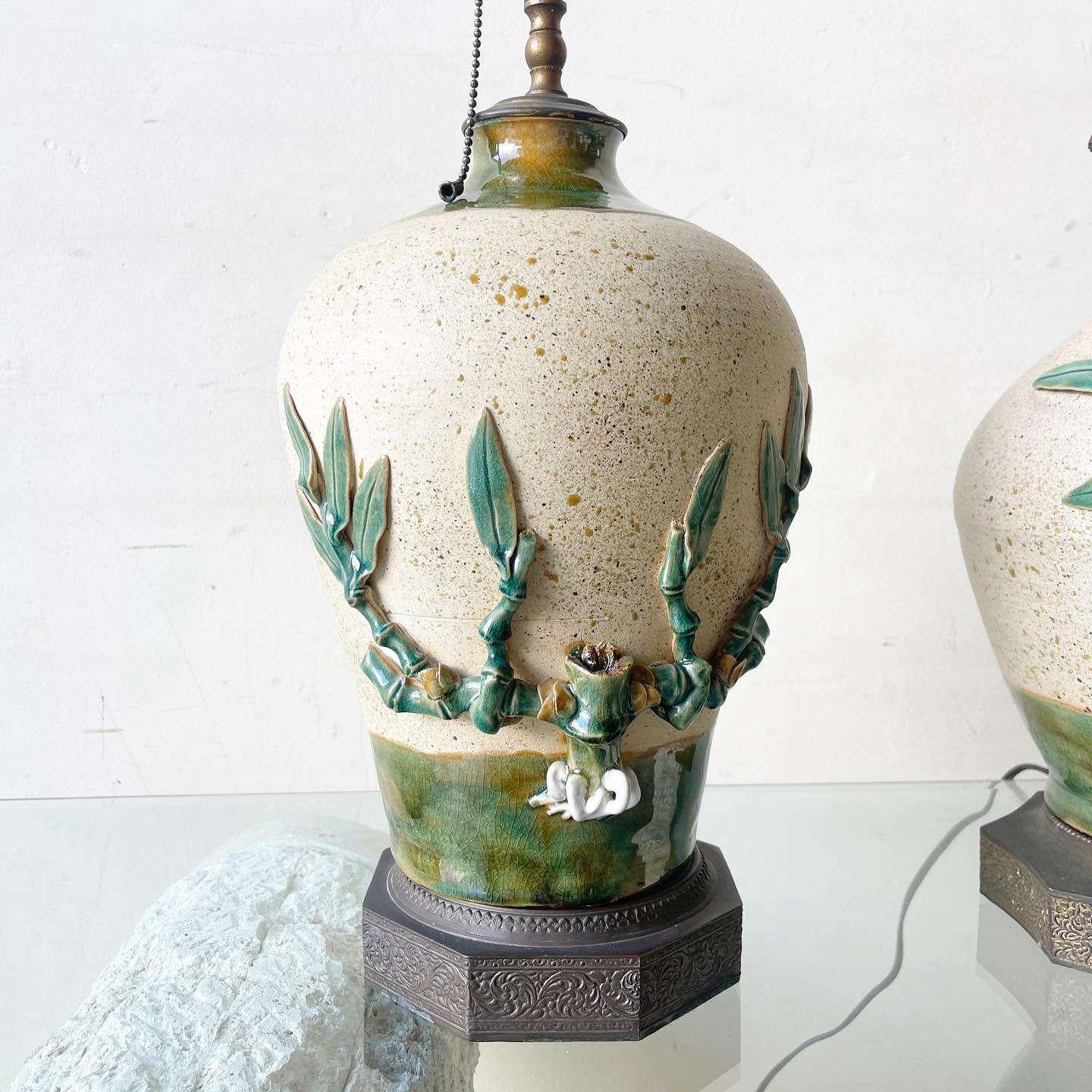 Amazing pair of vintage ceramic table lamps with brass brasses and tops. Each features a hand painted and sculpted bamboo plant growing around the sides. One of the lamps displays a bird.
