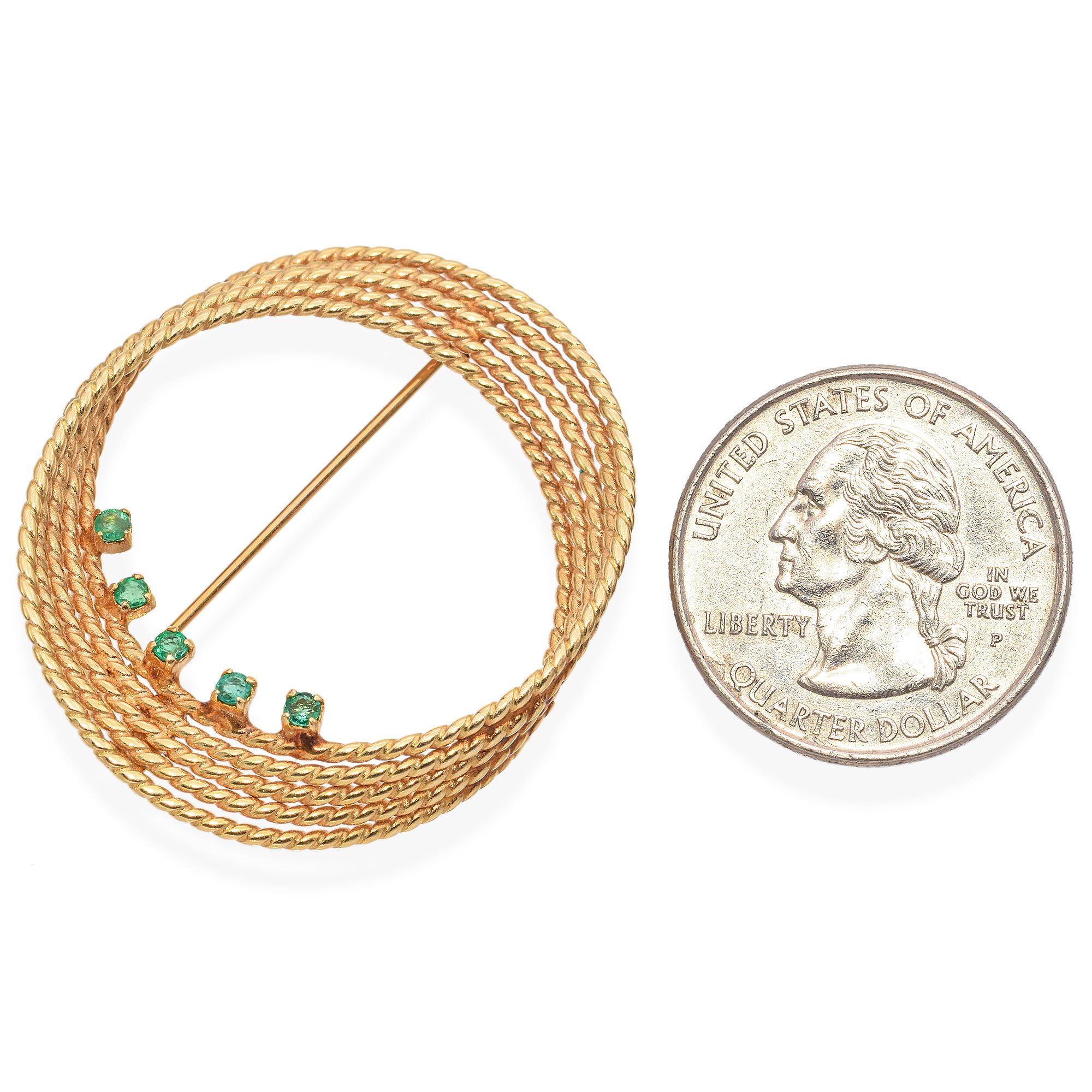 This exquisite piece features a circular motif crafted from yellow gold. The brooch showcases a series of twisted gold ropes, elegantly layered to form a captivating spiral pattern. Adorning the inner curve are five vibrant green emeralds, each