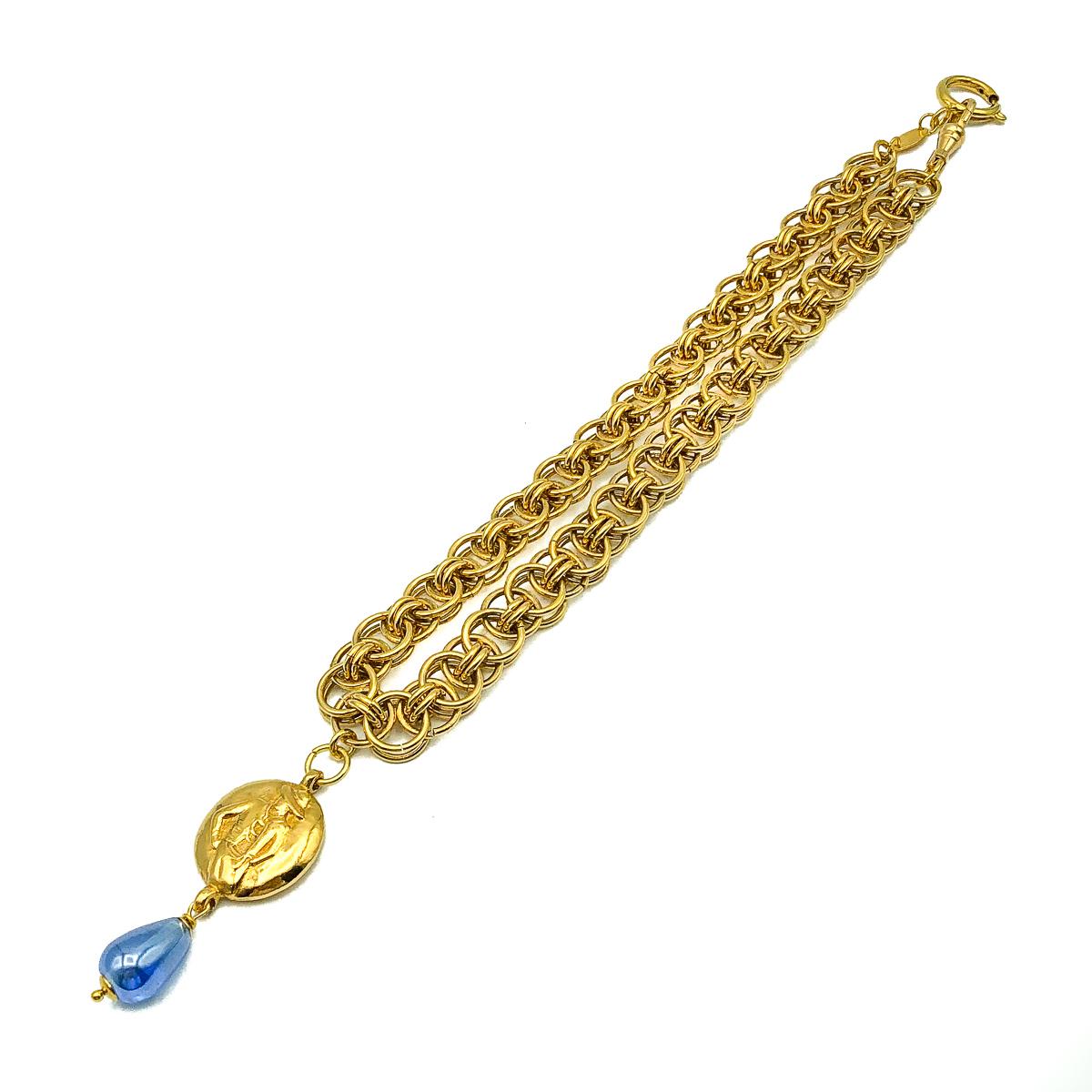 Un magnifique collier Coco vintage de Chanel. Réalisé en métal doré et en verre. Il s'agit d'une chaîne à anneau double avec un pendentif représentant Coco Chanel d'un côté et le nom CHANEL de l'autre. Fini avec une larme en verre bleu. En très bon