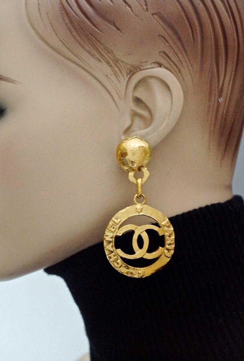 Vintage CHANEL Cutout Logo Medallion Hammered Dangling Earrings

Measurements: 
Height: 3 inches (7.8 cm)
Width: 1.57 inches (4 cm)
Weight per Earring: 14 grams

Features:
- 100% Authentic CHANEL.
- Hammered cutout Chanel CC logo medallion.
-