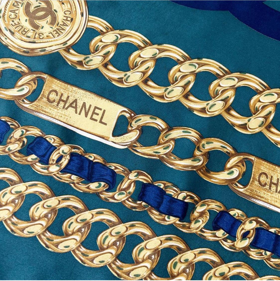 Cette écharpe vintage Chanel est confectionnée en 100 % soie vert émeraude et présente les maillons de chaîne dorés emblématiques de Chanel ainsi qu'un CC bleu marine au centre et des bords roulés cousus à la main. Mesure 34 x 33 pouces. Fabriquées