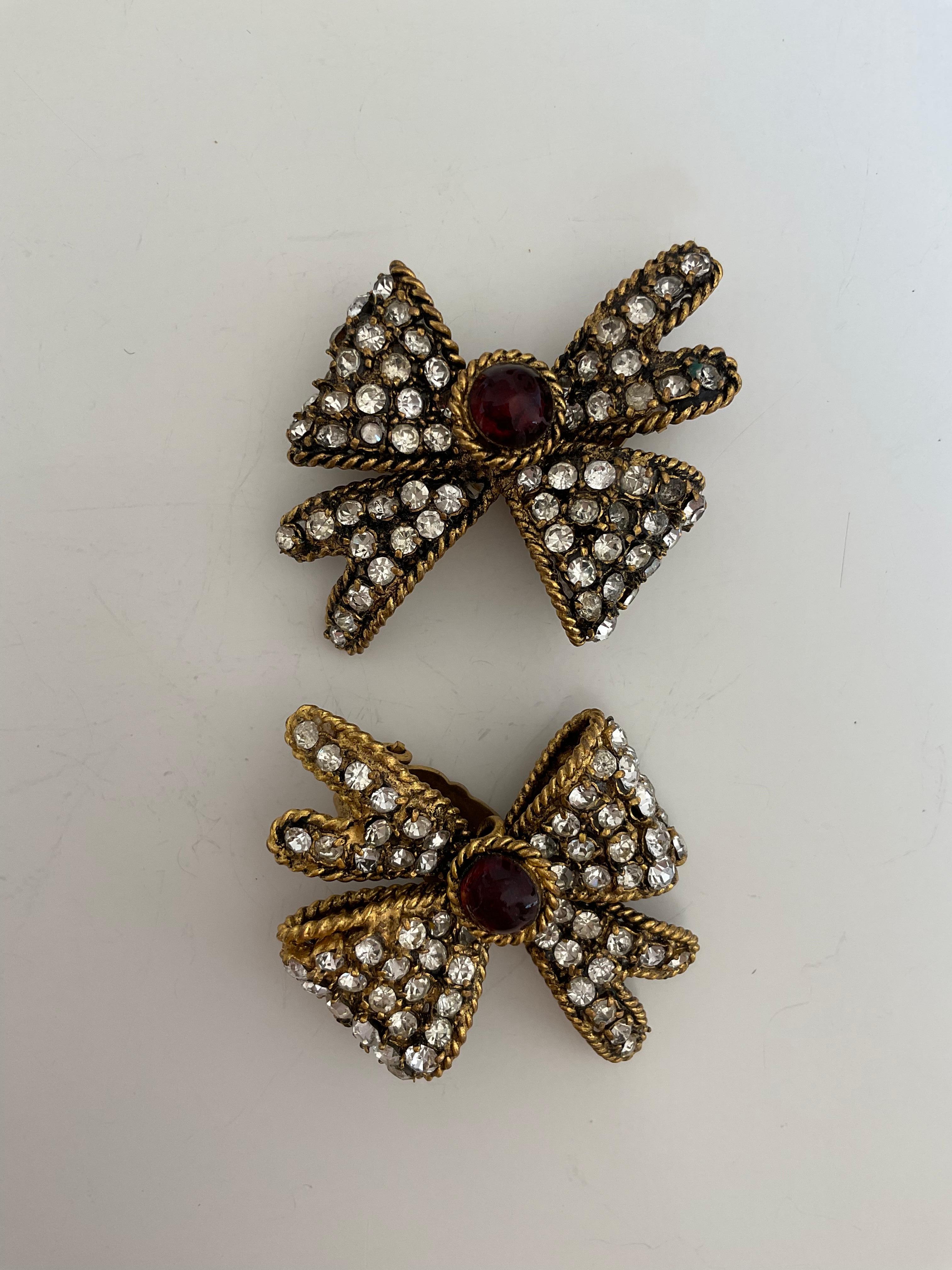 This pair of earrings are rare Vintage from the House of CHANEL from the 80's. Designed like dainty bows, those clip-on earrings are gold plated, encrusted with rhinestones and a round dark red stone in “pate de verre” at the centre, where the nods