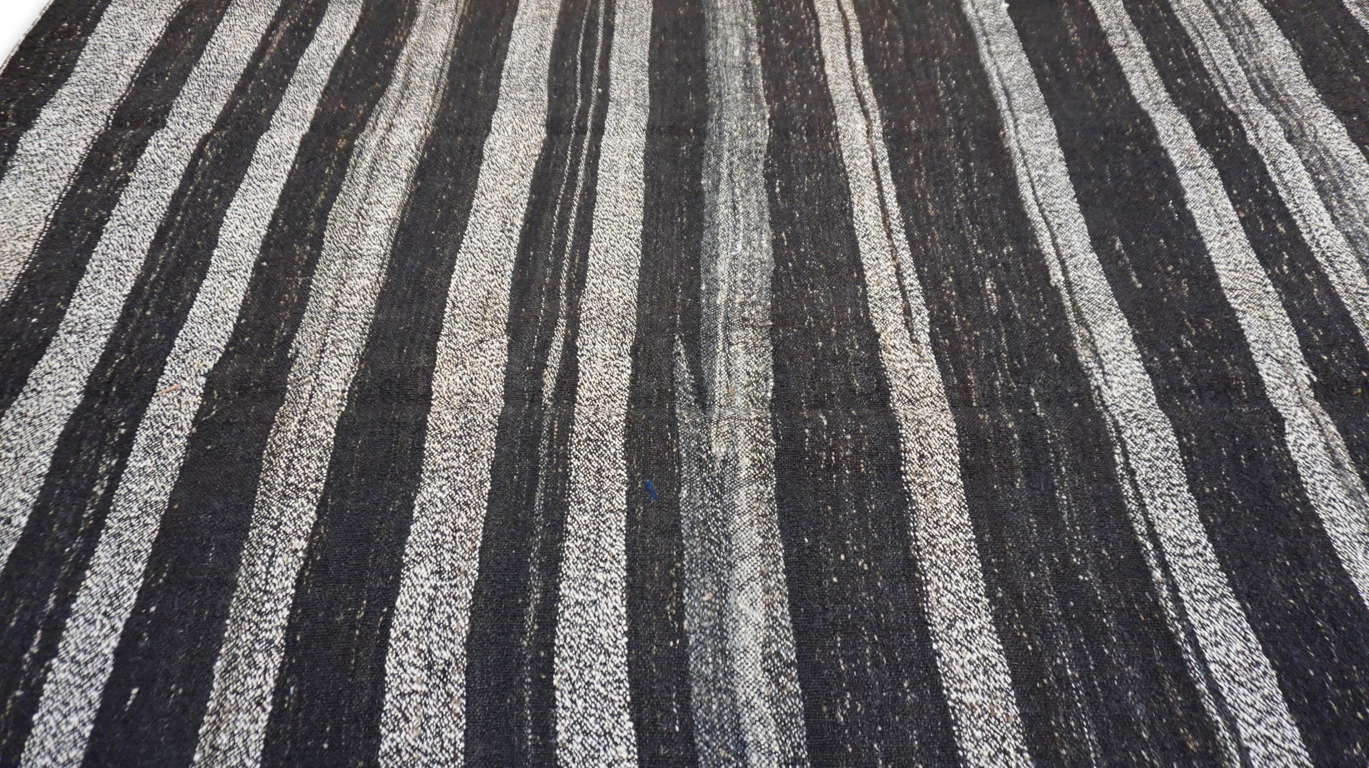 Questo  Il kilim in lana/pelo di capra presenta un'estetica monocromatica a strisce color carbone, grigio, marrone e argento, che incarna sia l'eredità tribale che il minimalismo moderno. Le strisce organiche e irregolari creano un ritmo visivo