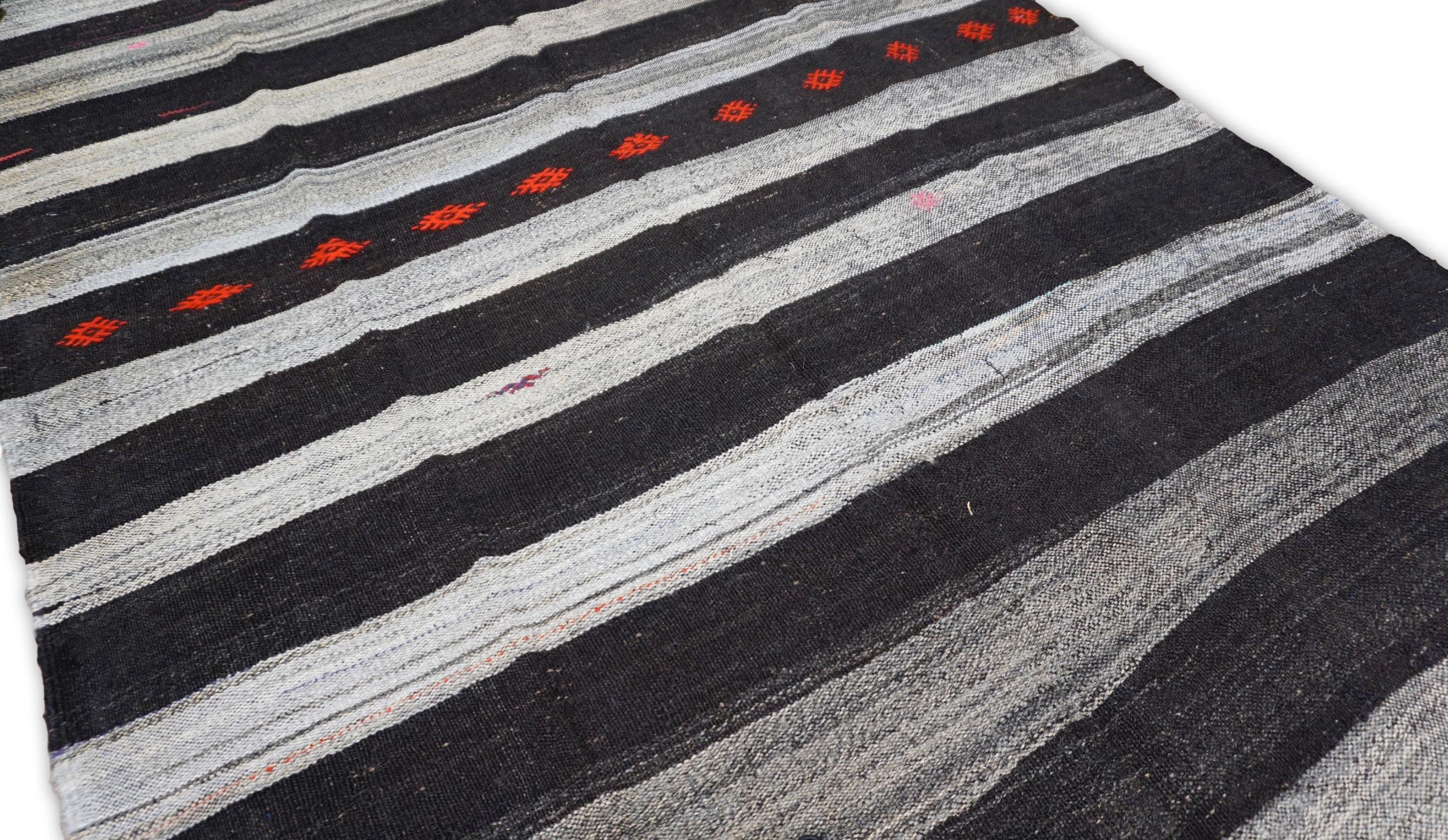 This wool kilim rug is a stunning example of traditional craftsmanship blended with bold, modern aesthetics. Featuring horizontal stripes in varying shades of charcoal, red, and gray, the rug creates a textured visual effect that brings depth and