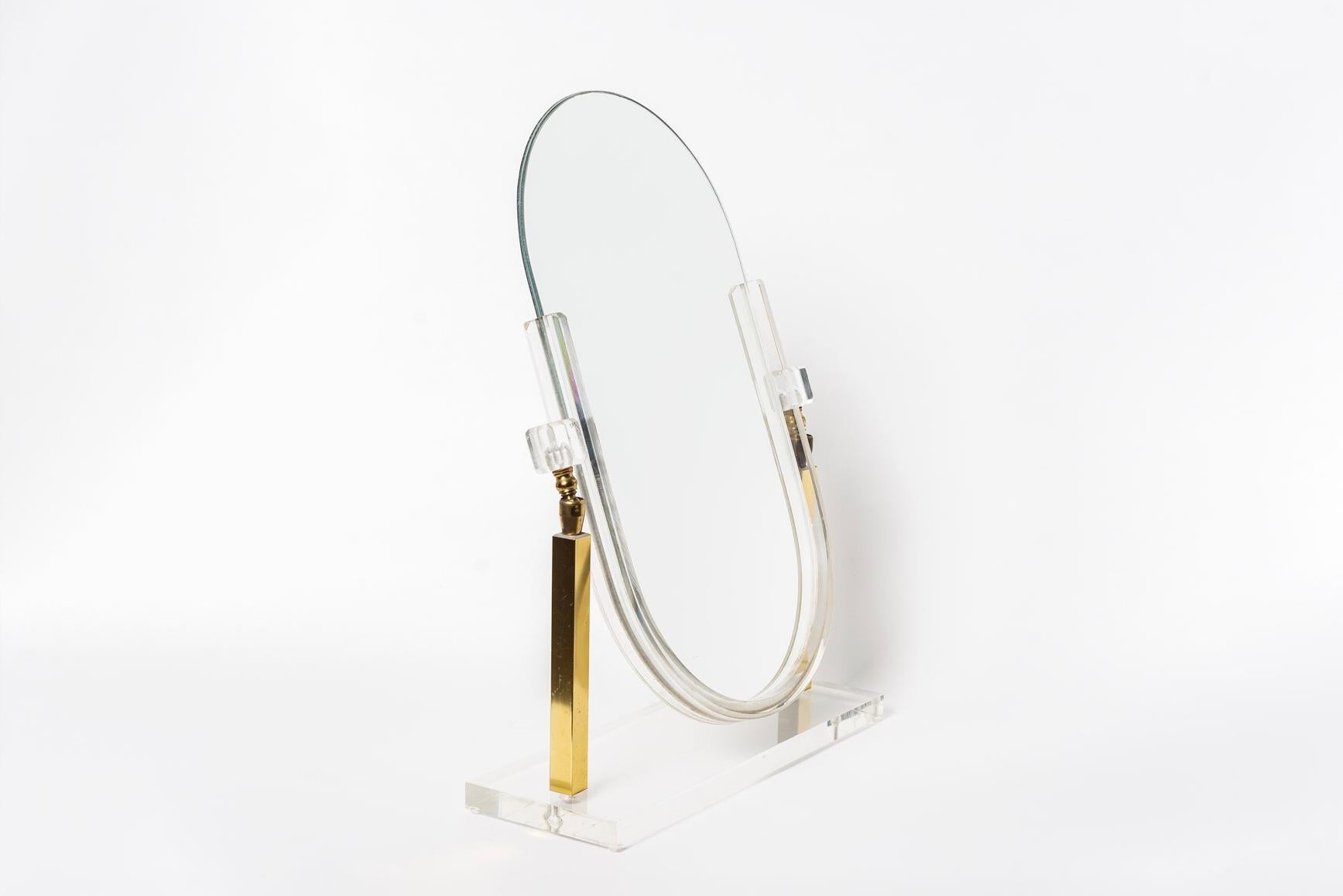 This vintage Mid-Century Modern brass and Lucite vanity mirror in the style of Charles Hollis Jones is circa 1970. The elegant Minimalist design features clean geometric lines; with a frameless oval mirror held suspended from two brass pedestal arms