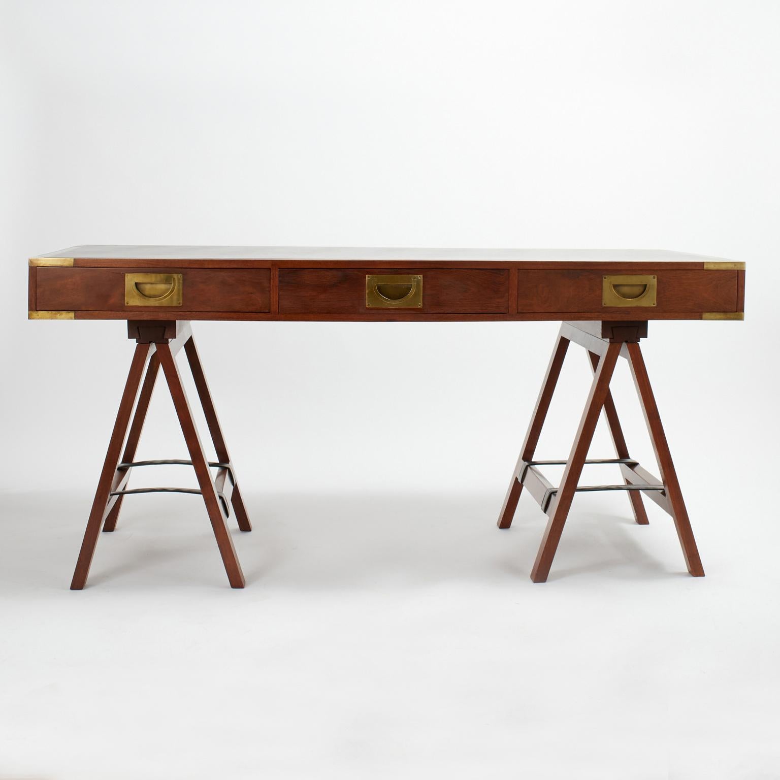 Dashing mid century campaign style desk crafted in teak with a black leather top banded in brass having a case with a fold down center drawer and campaign style brass hardware sitting on removable folding sawhorse legs with leather straps. Signed
