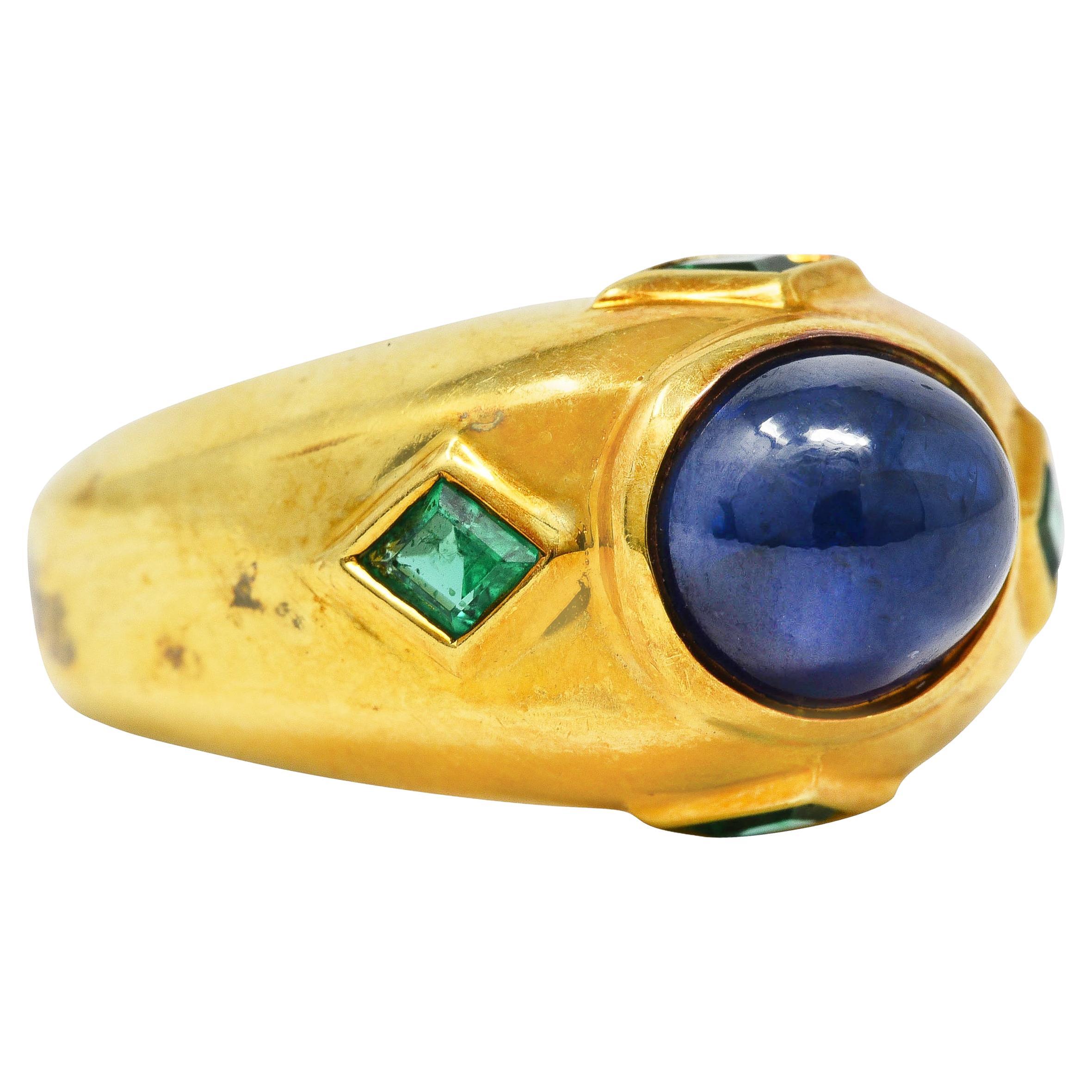 Yellow gold bombè band centers a sapphire cabochon weighing approximately 4.85 carat. Blue in color and semi-transparent with natural inclusions while exhibiting mild chatoyancy. Bezel set in a polished gold surround with navette cut emerald accents