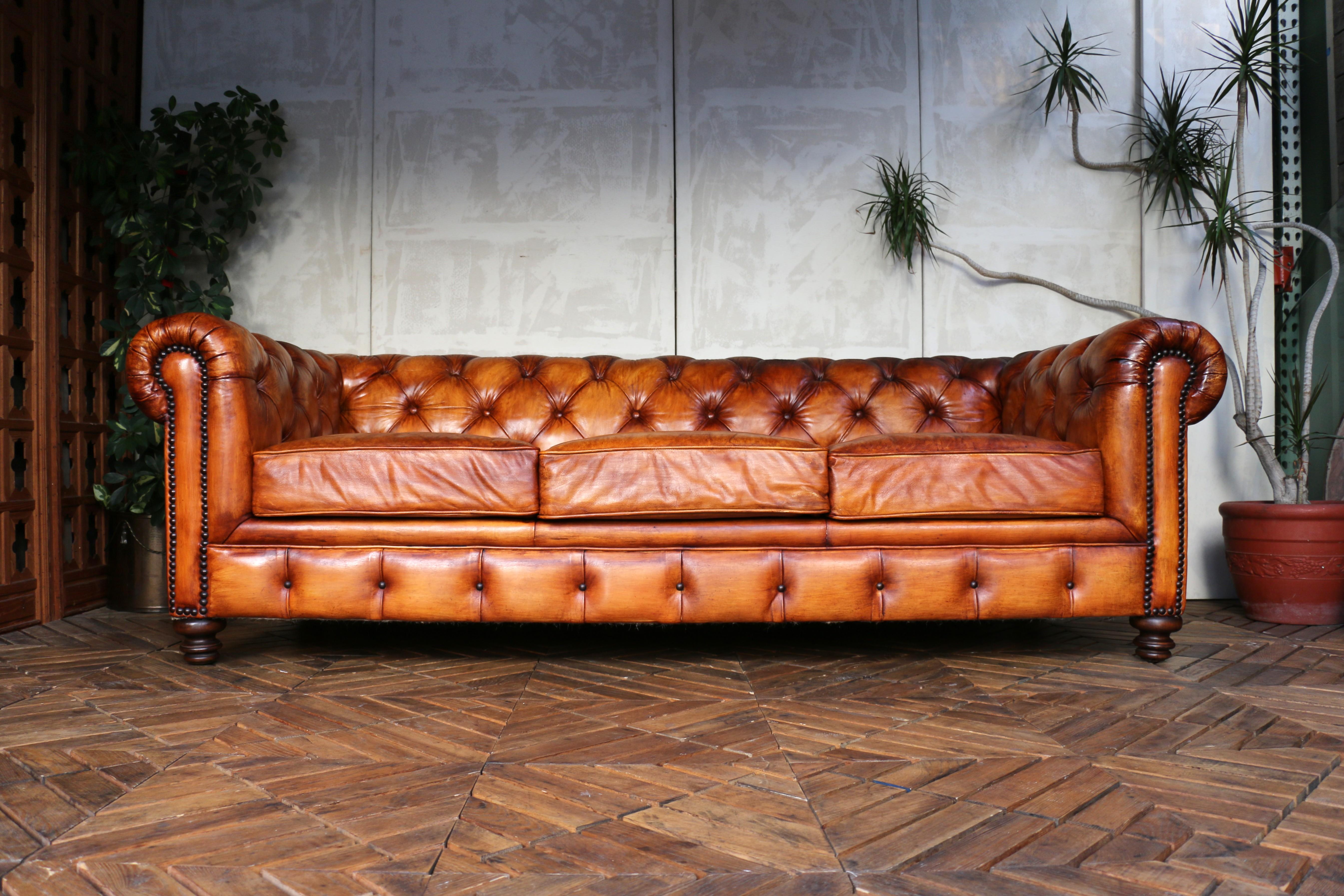 Un impresionante sofá vintage de estilo Chesterfield, tapizado en cuero marrón envejecido. Esta pieza clásica luce el característico respaldo acolchado con botones y los brazos enrollados, que destilan una elegancia británica atemporal. El cuero