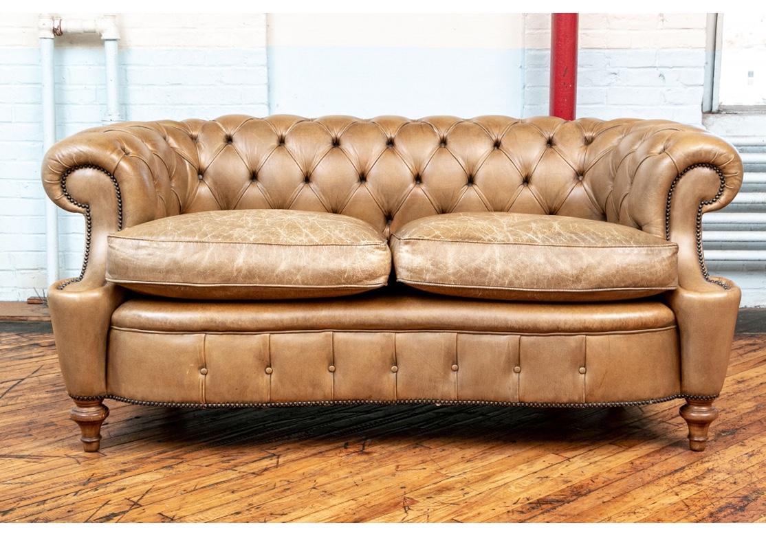 A two seat shaped sofa in tan leather with button tufted rolled back. Rolled sloping arms into the rolled supports. The shaped skirt with button tufting. Nail- head trim on the arms, skirt and back. Raised on turned dark wood legs front and back.