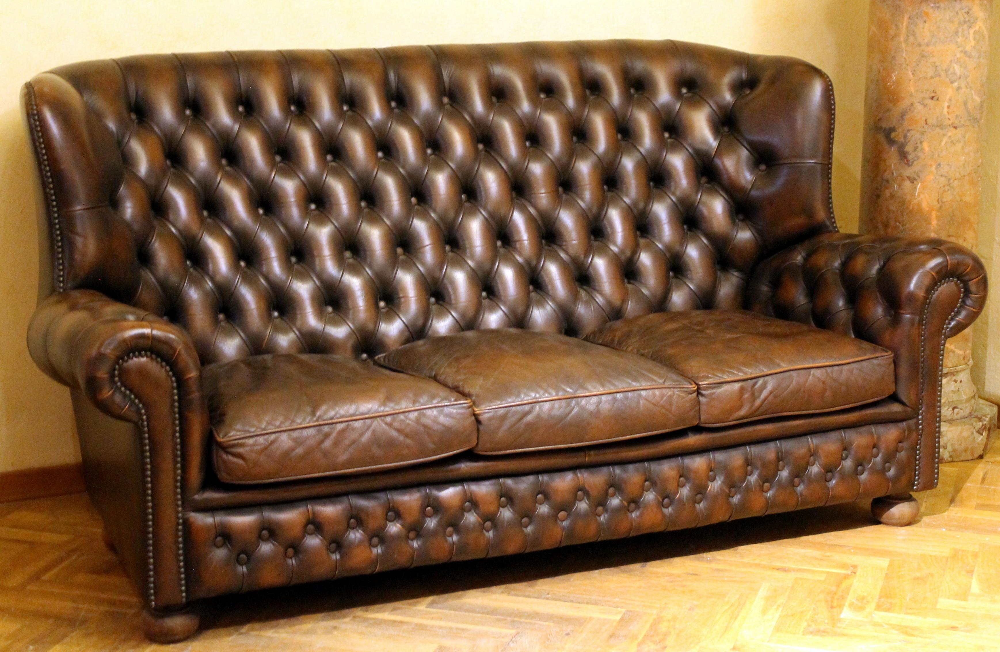 This English vintage brown leather Chesterfield sofa with button trufted back and arms offers three seats and high back, striking profile thanks to slight winged back, scrolled arms and gently scalloped back rail, the all resting on solid turned