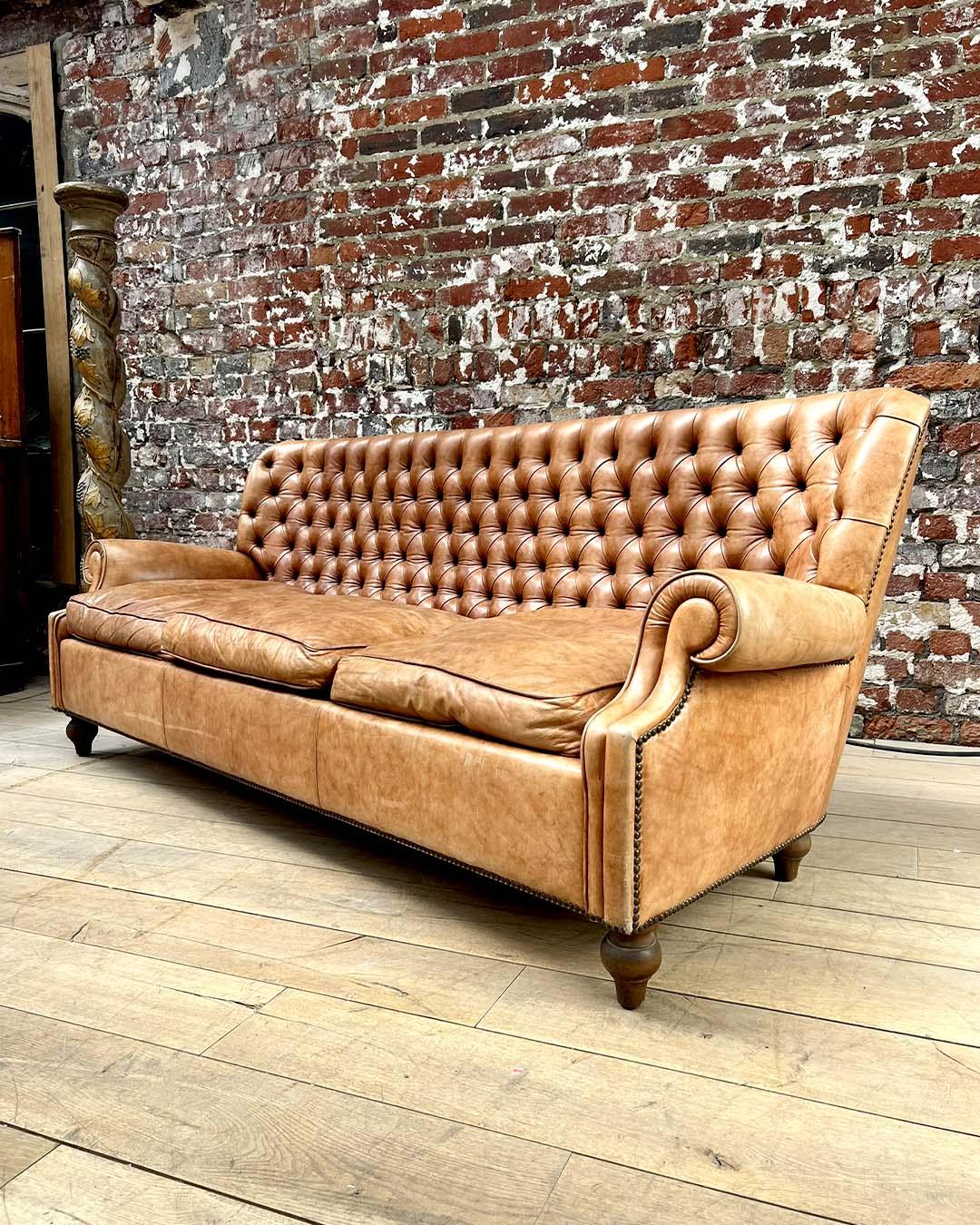 A handsome and generously sized Chesterfield-style sofa upholstered in rich, naturally patinated tan leather. The deep-buttoned backrest showcases classic tufting, while the elegant scroll arms are accentuated with brass nailhead trim. Raised on