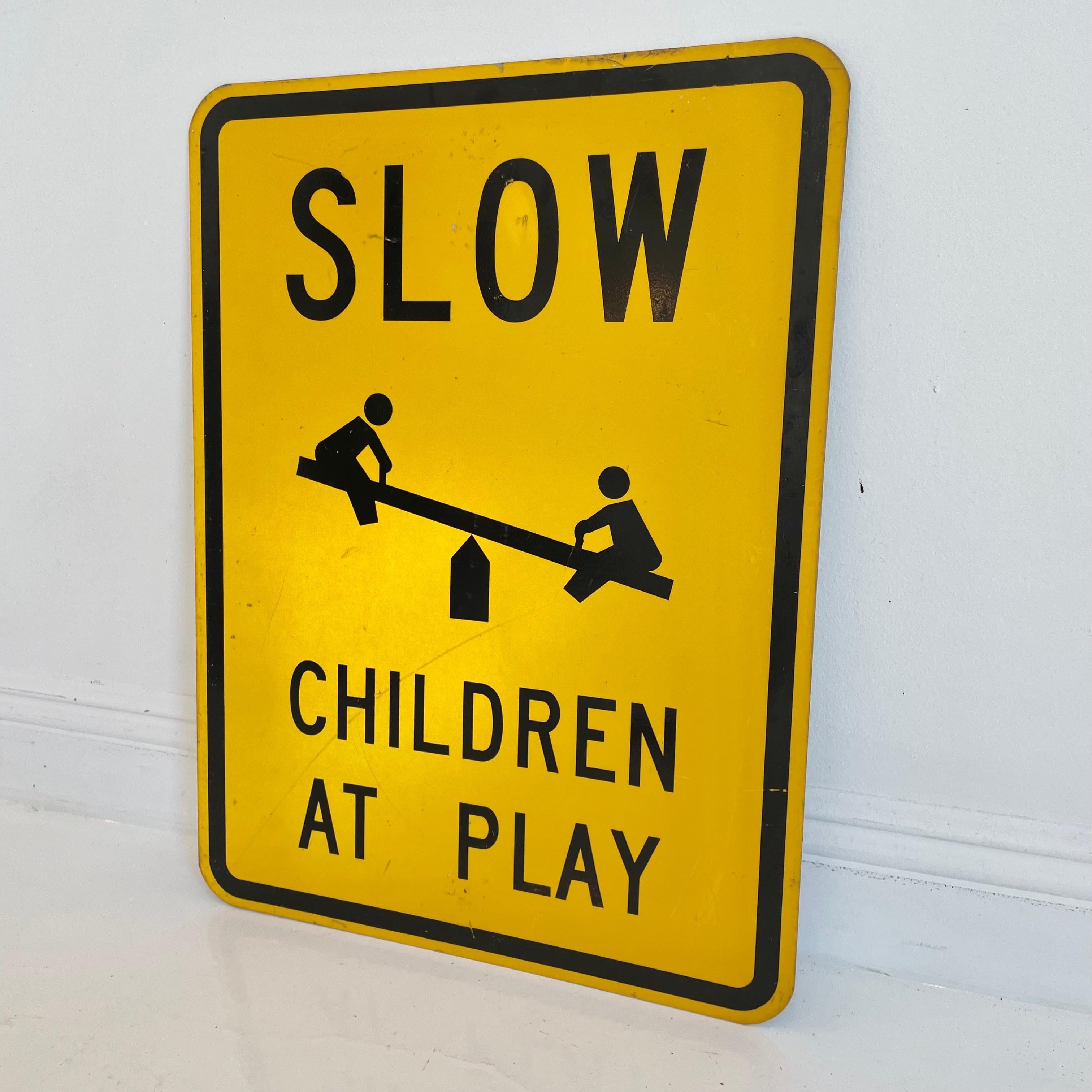 Unique vintage street sign that reads 'SLOW - Children at Play' Black lettering on a reflective yellow background and black trim. Fun graphics depicting two children on a teeter totter. Great vintage condition. Classic coloring.