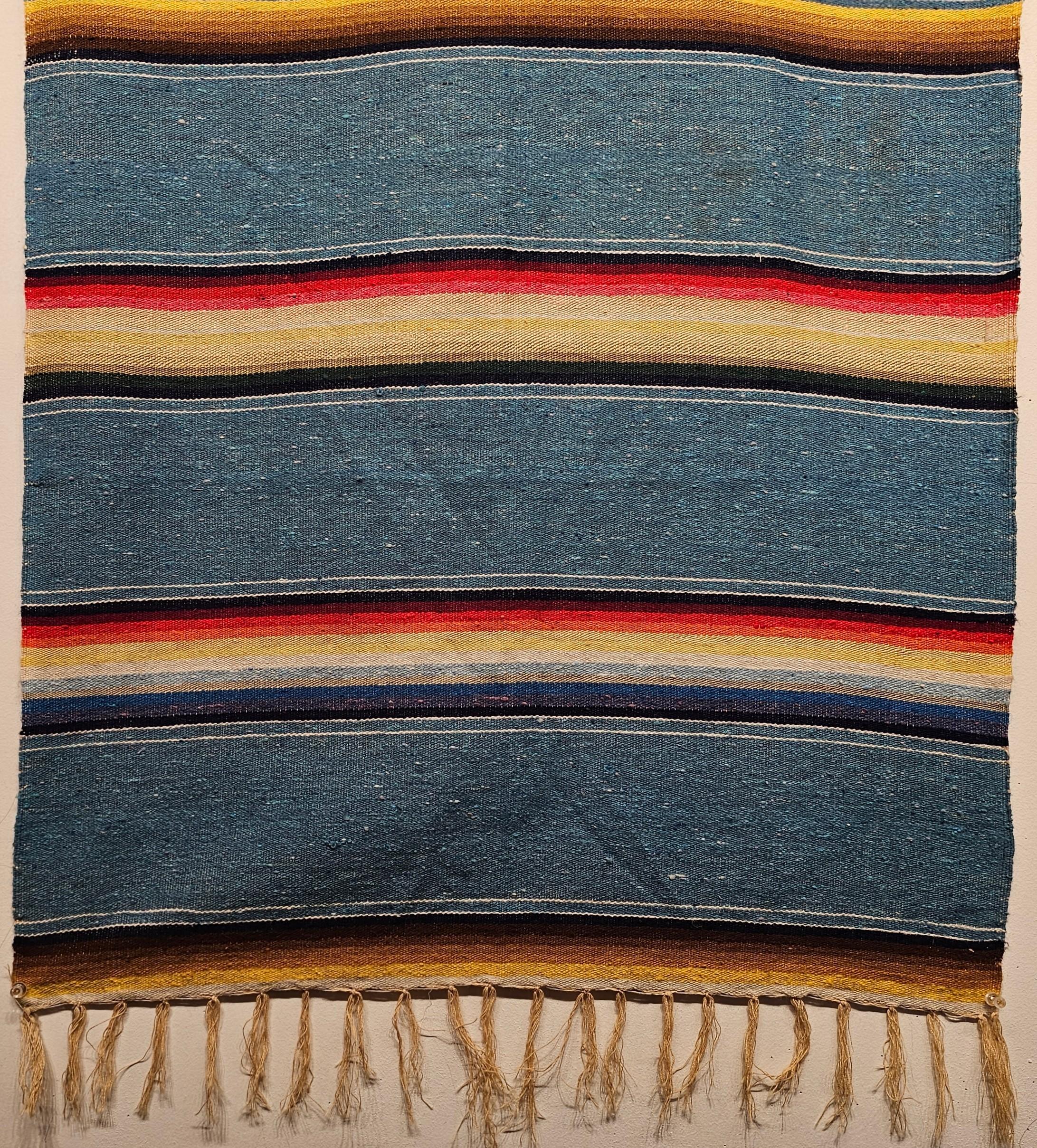 A beautiful Chimayo rug was woven in Mexico in the 2nd quarter of the 1900s. The rug has a beautiful baby blue color with stripes in brilliant colors including blue, brown, yellow, black, and cream throughout. The rug is extremely finely woven and