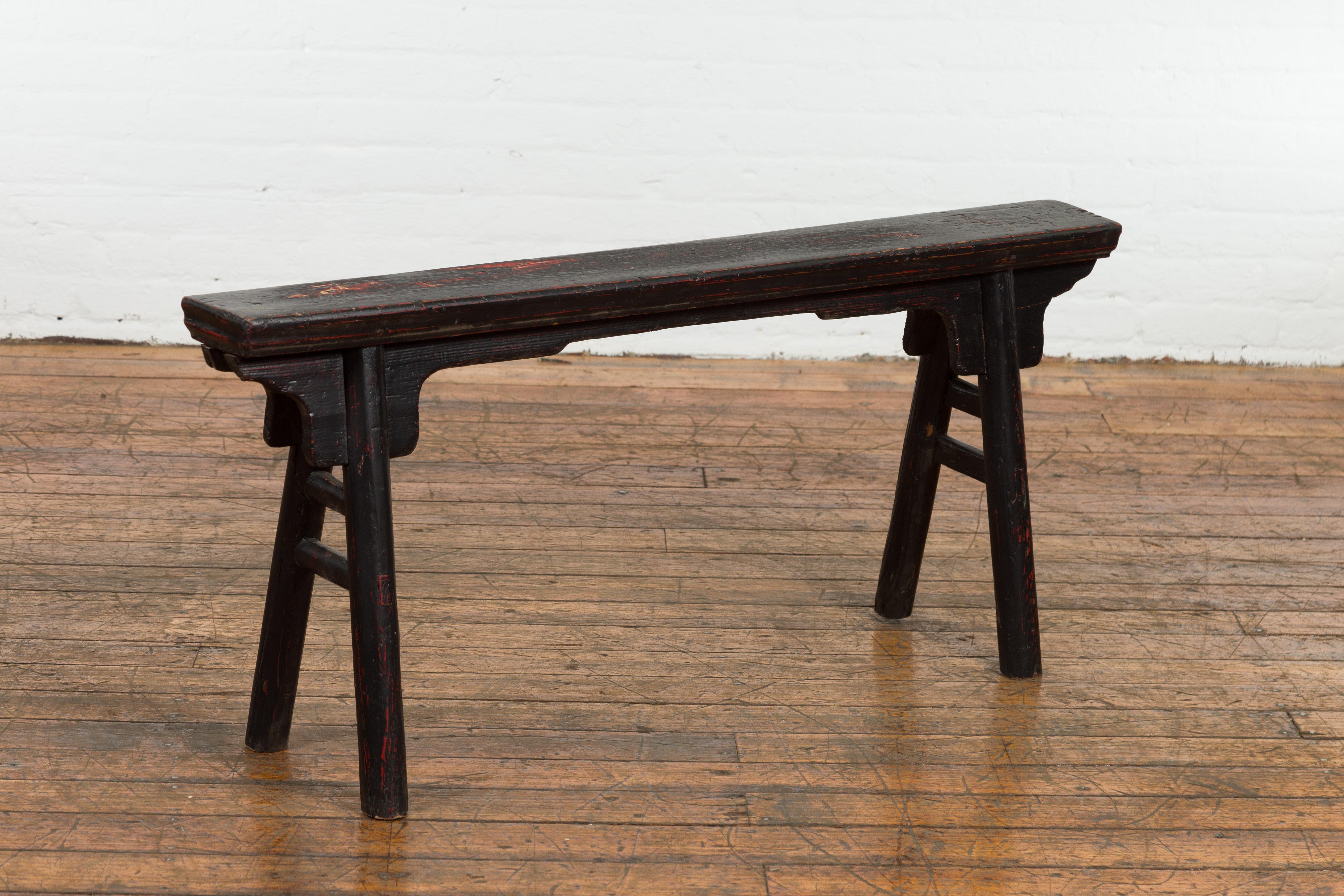 A vintage Chinese A-Frame black painted bench from the mid 20th century with red color coming through, nicely distressed patina, splaying legs, carved spandrels and double side stretchers. Revel in the rustic charm and charming design of this