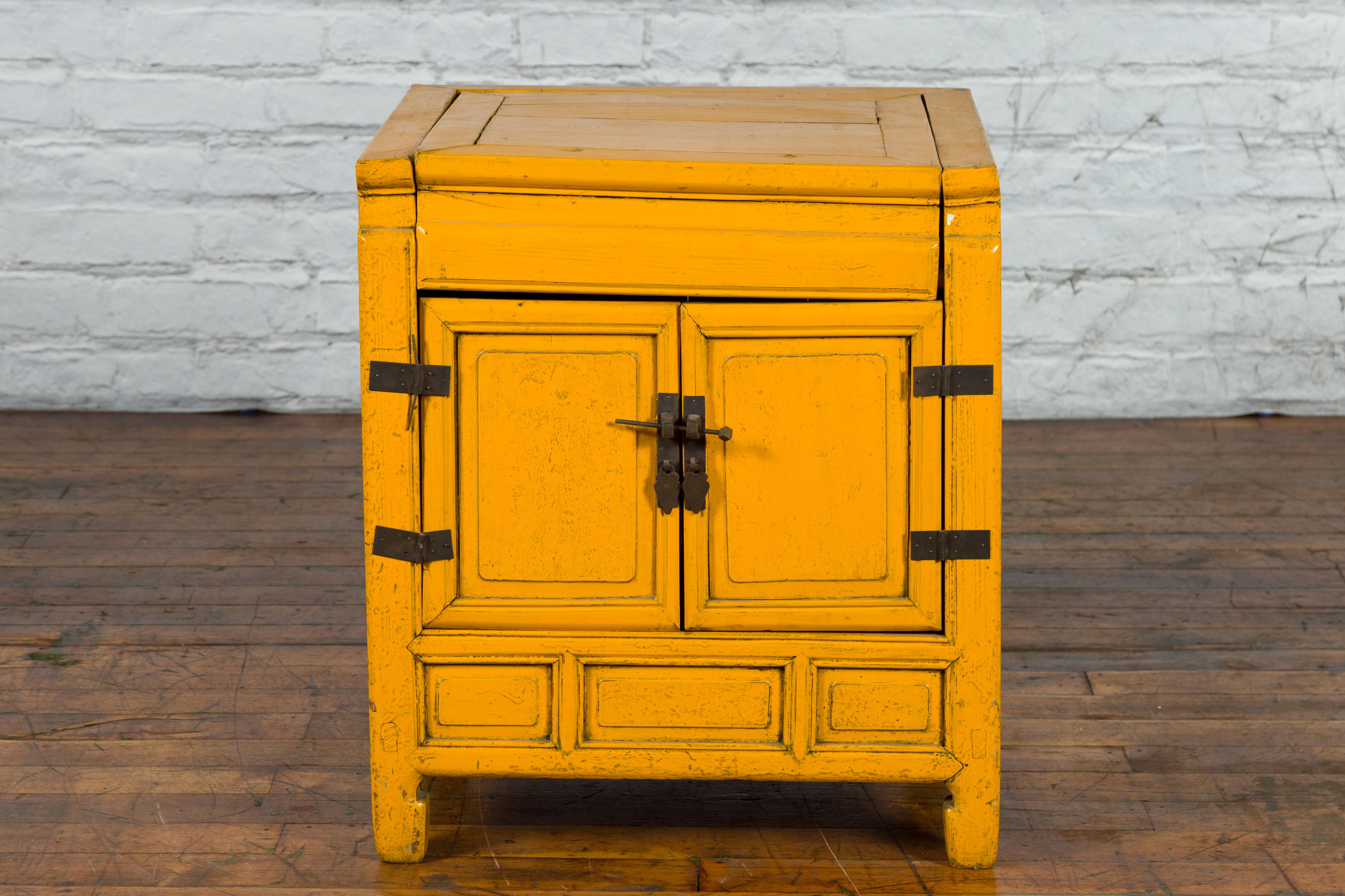 A vintage Chinese bedside cabinet from the mid 20th century, with distressed yellow lacquer, lift top, two doors, brass hardware and horse hoof feet. Created in China during the Midcentury period, this bedside cabinet features a linear shape