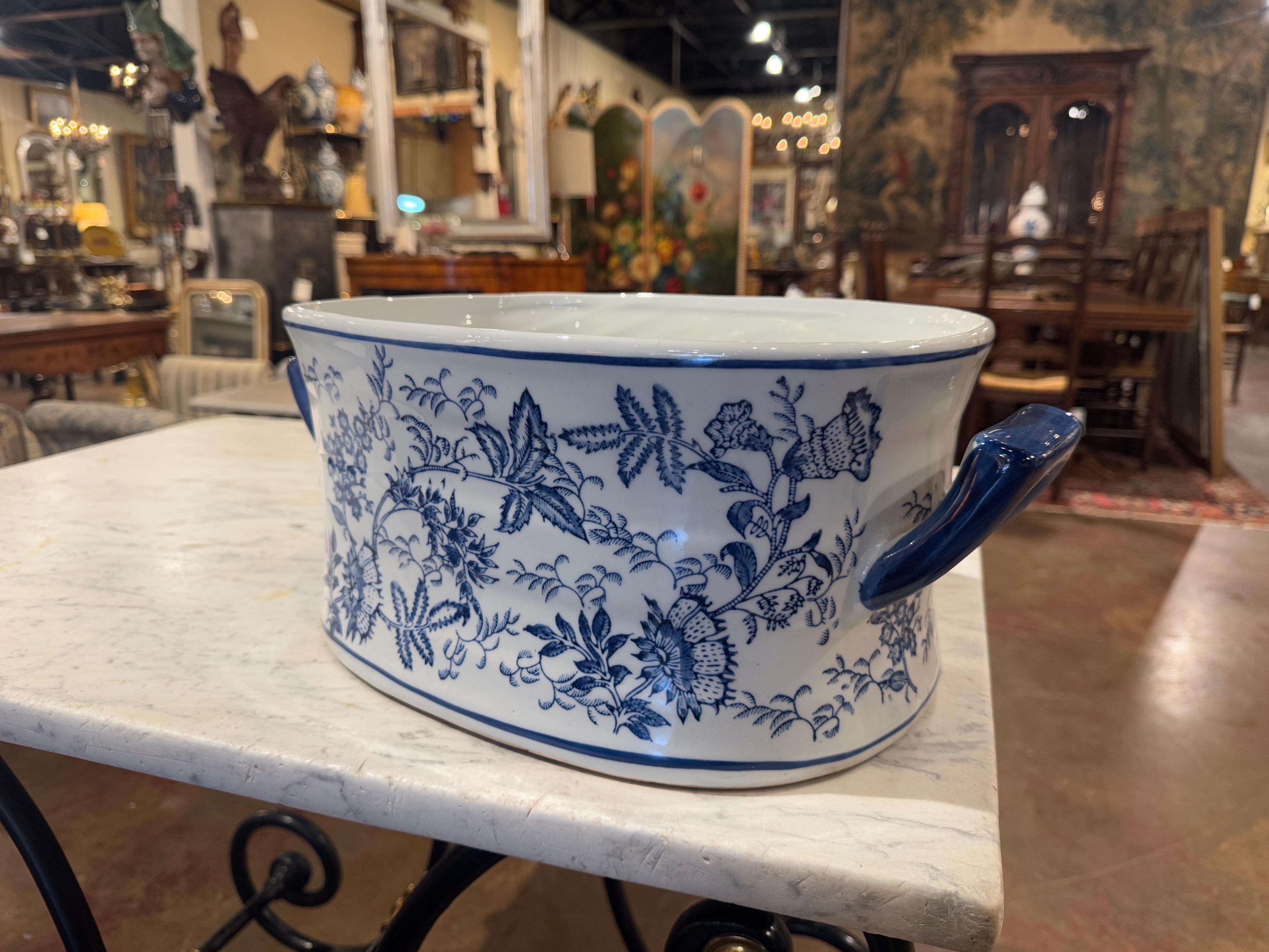 Crafted in China circa 1990 and oval in shape, the vintage porcelain foot bath is dressed with two side handles; it is decorated with hand painted floral and foliage motifs in a blue and white palette. This elegant and colorful jardinière is in