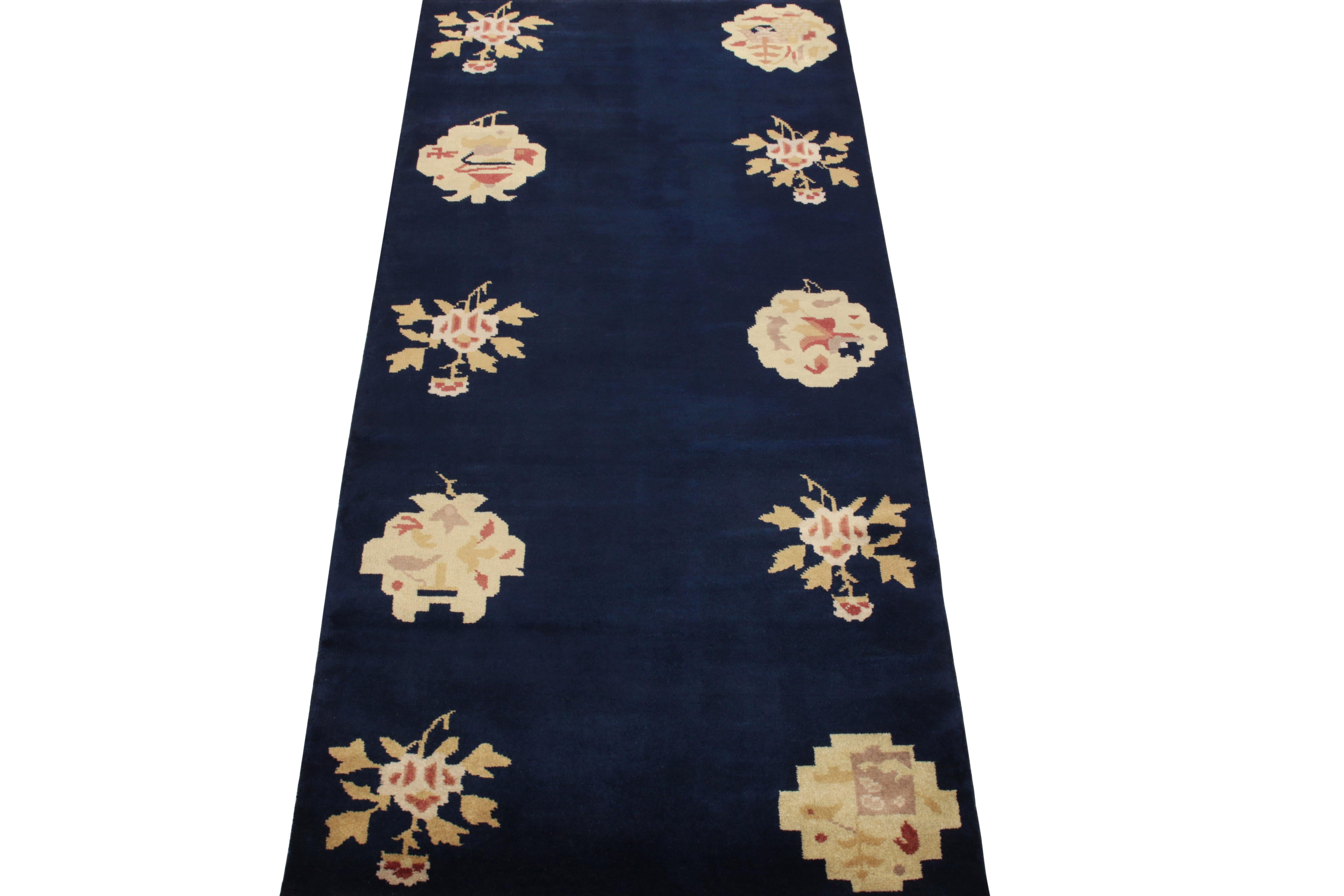 Hand-knotted in wool, this 3x6 vintage ode to Chinese art deco rugs of the 1920s features a series of alternating floral designs in subtle golden-beige, carrot red with light purple on a lush blue backdrop carrying light-dark spots further