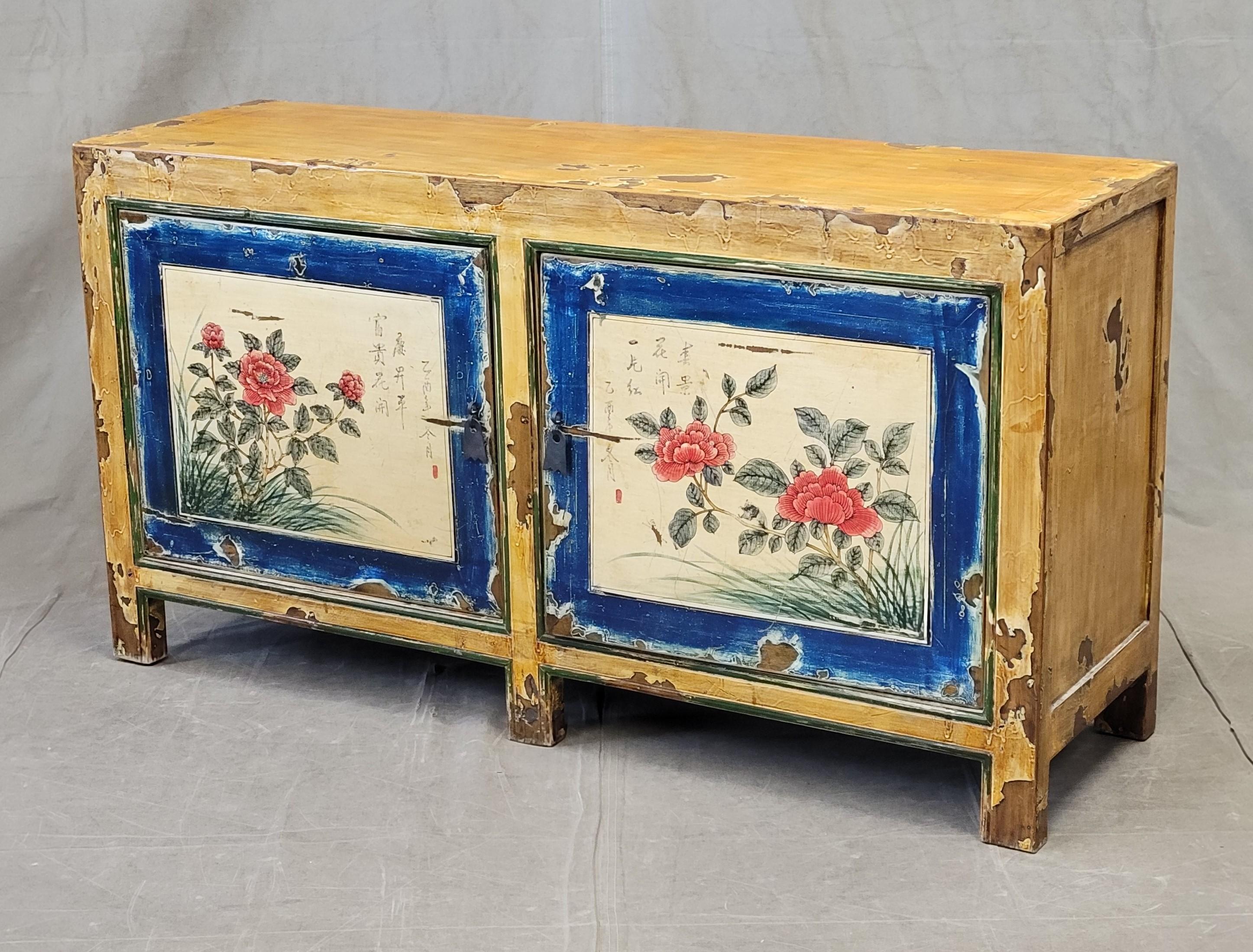 Una encantadora consola vintage de laca china en preciosos tonos ocres, azules, blancos, rojos y verdes. Los paneles florales pintados de las puertas están acentuados con escritura china. Observa los desgastes en el acabado lacado, ver fotos, que
