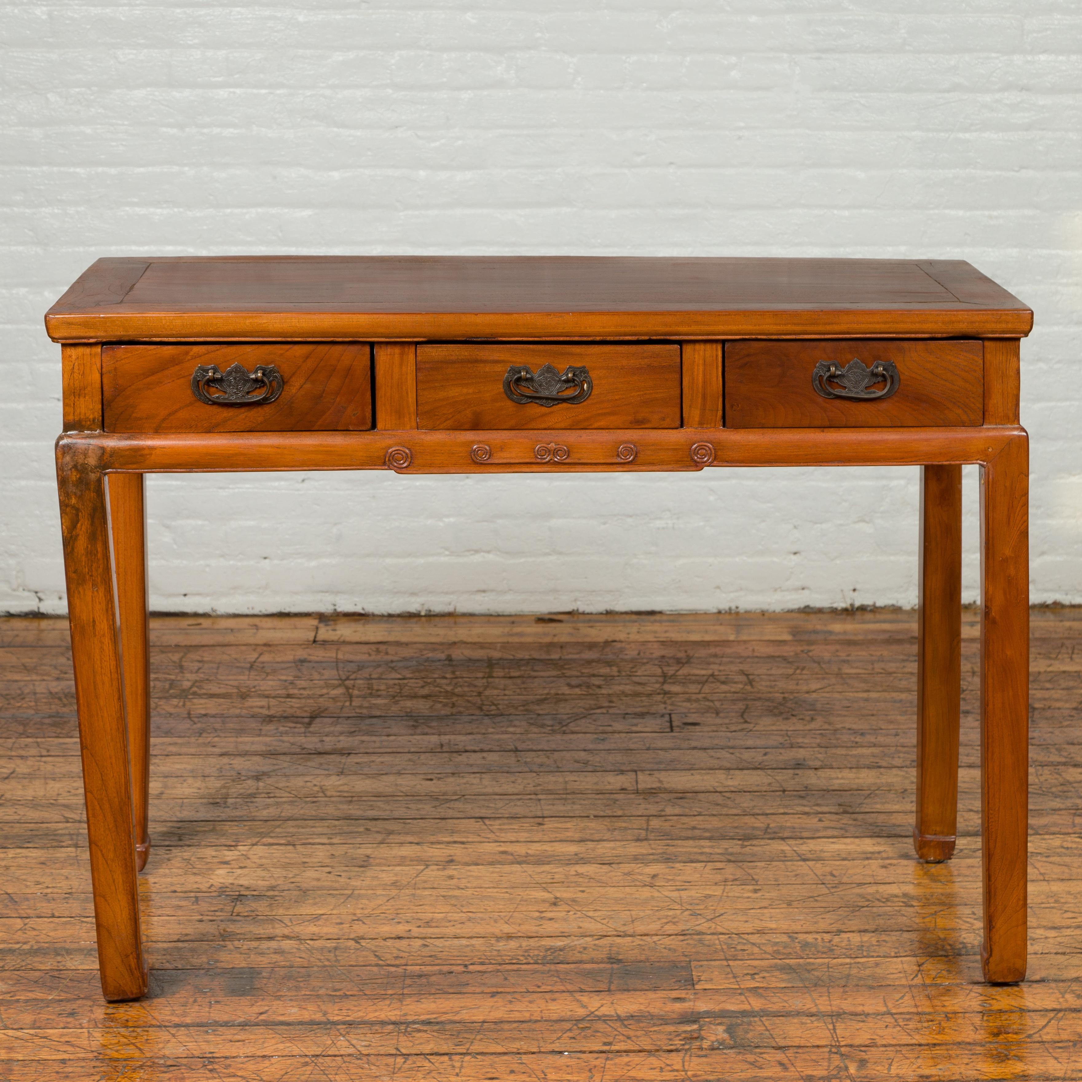A vintage Chinese elmwood desk from the mid-20th century, with three drawers and petite swirling scrolls. Born in China during the midcentury period, this elm writing table features a rectangular top sitting above three drawers, each fitted with