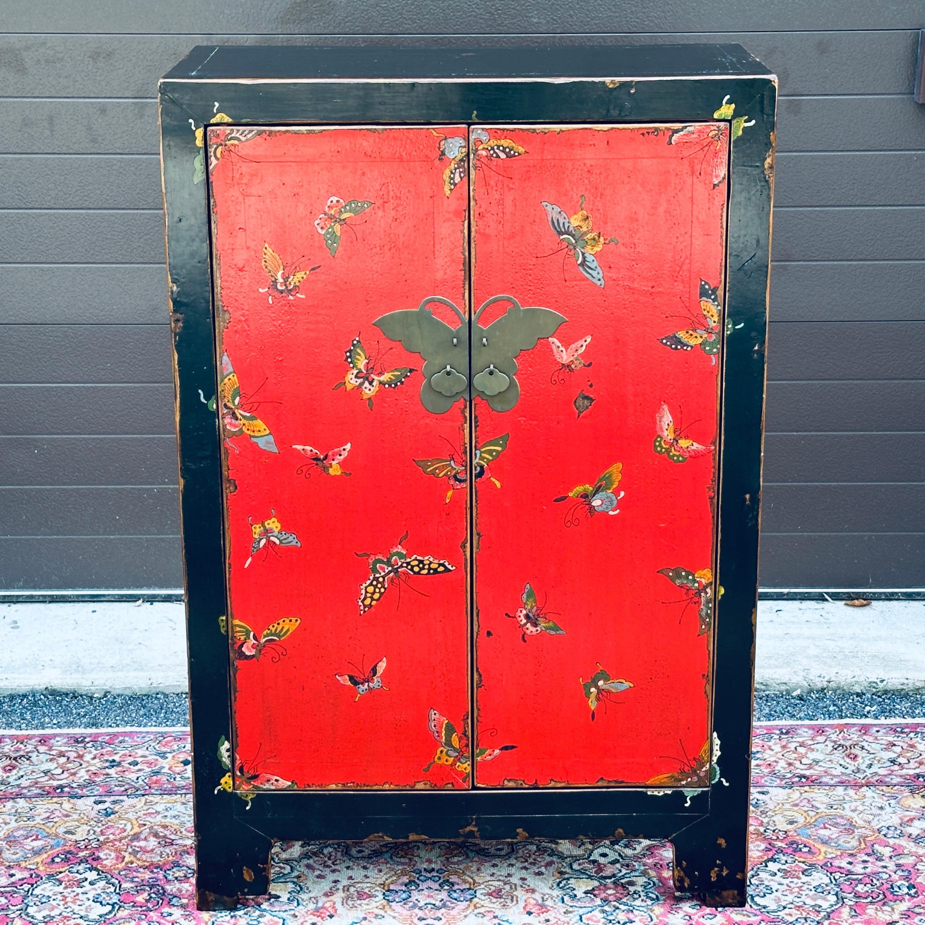 Mueble chino vintage lacado en rojo y negro con detalle de mariposa pintada a mano y herrajes de mariposa de latón. Dos puertas de longitud completa se abren para revelar el espacio del armario y dos estantes extraíbles. Se trata de una reproducción