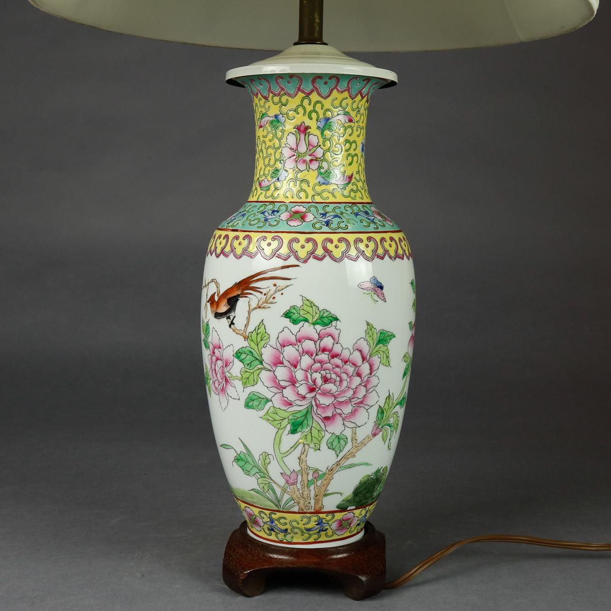 A vintage Chinese table lamp offers lidded porcelain vase in hourglass form with hand painted garden scene of flowers and birds, collar and base with repeating stylized foliate and scroll pattern bands, seated on Harwood base with scroll form feet,
