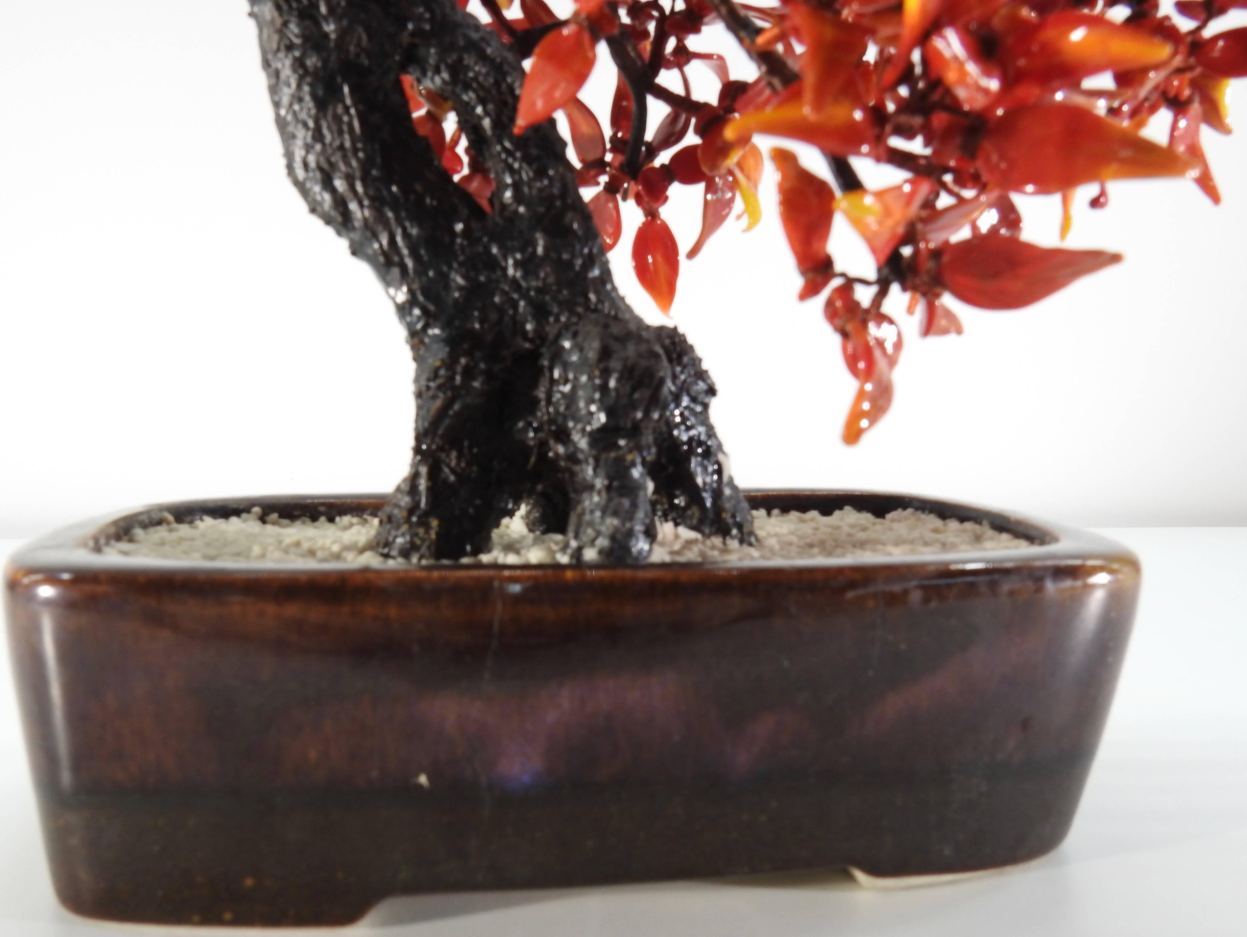 Burnished rust tones of polished glass form the leaves on this Bonsai tree from the 1940s. The branches are neatly covered in silk threads, coming out from the trunk. The tree is planted in a bed of tiny stones which is in a ceramic planter.
 