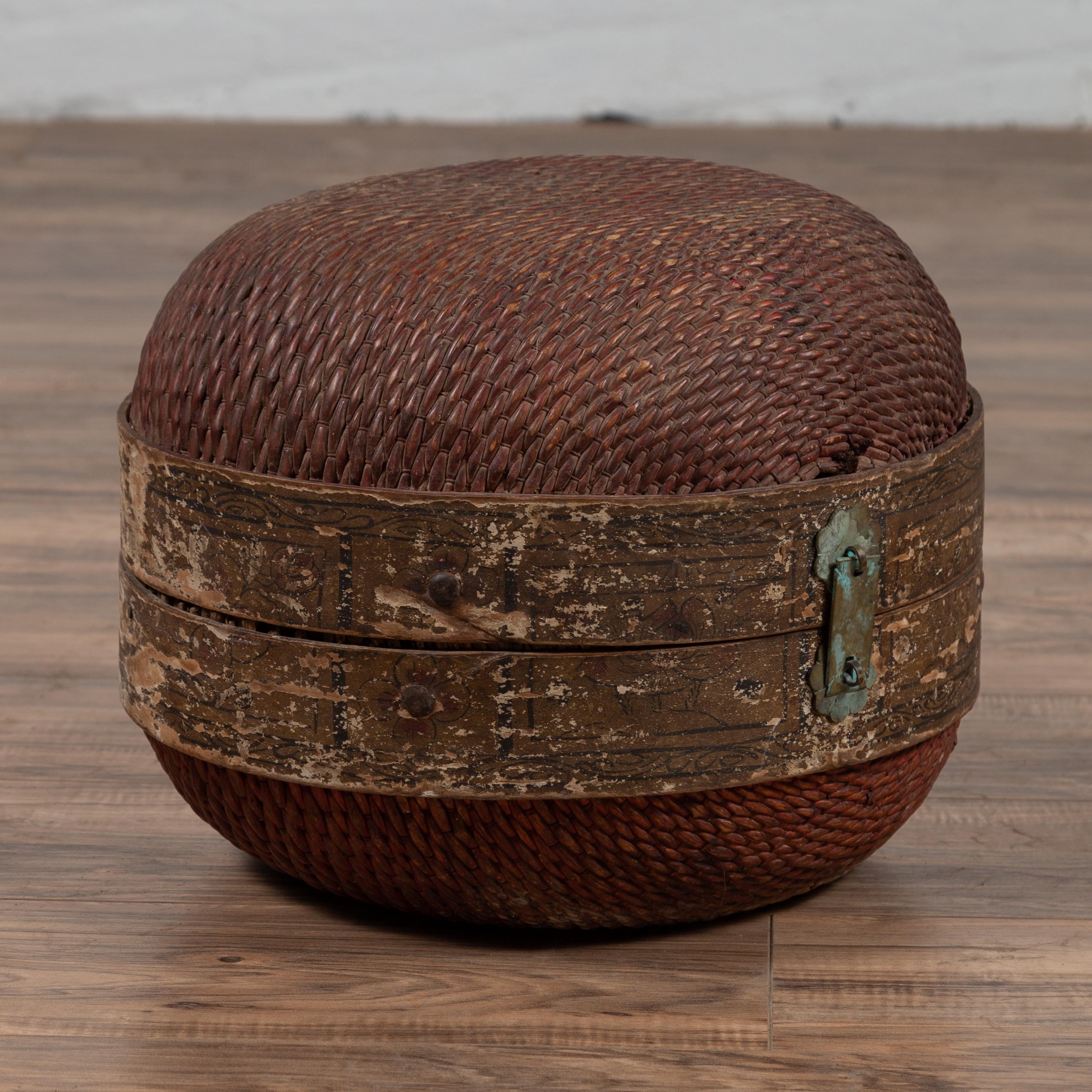 A vintage Chinese rattan hat box from the mid-20th century with weathered patina and oxidized accents. We have similar ones available. Born in China during the midcentury period, this rustic hat box features a rattan body, secured in the middle by