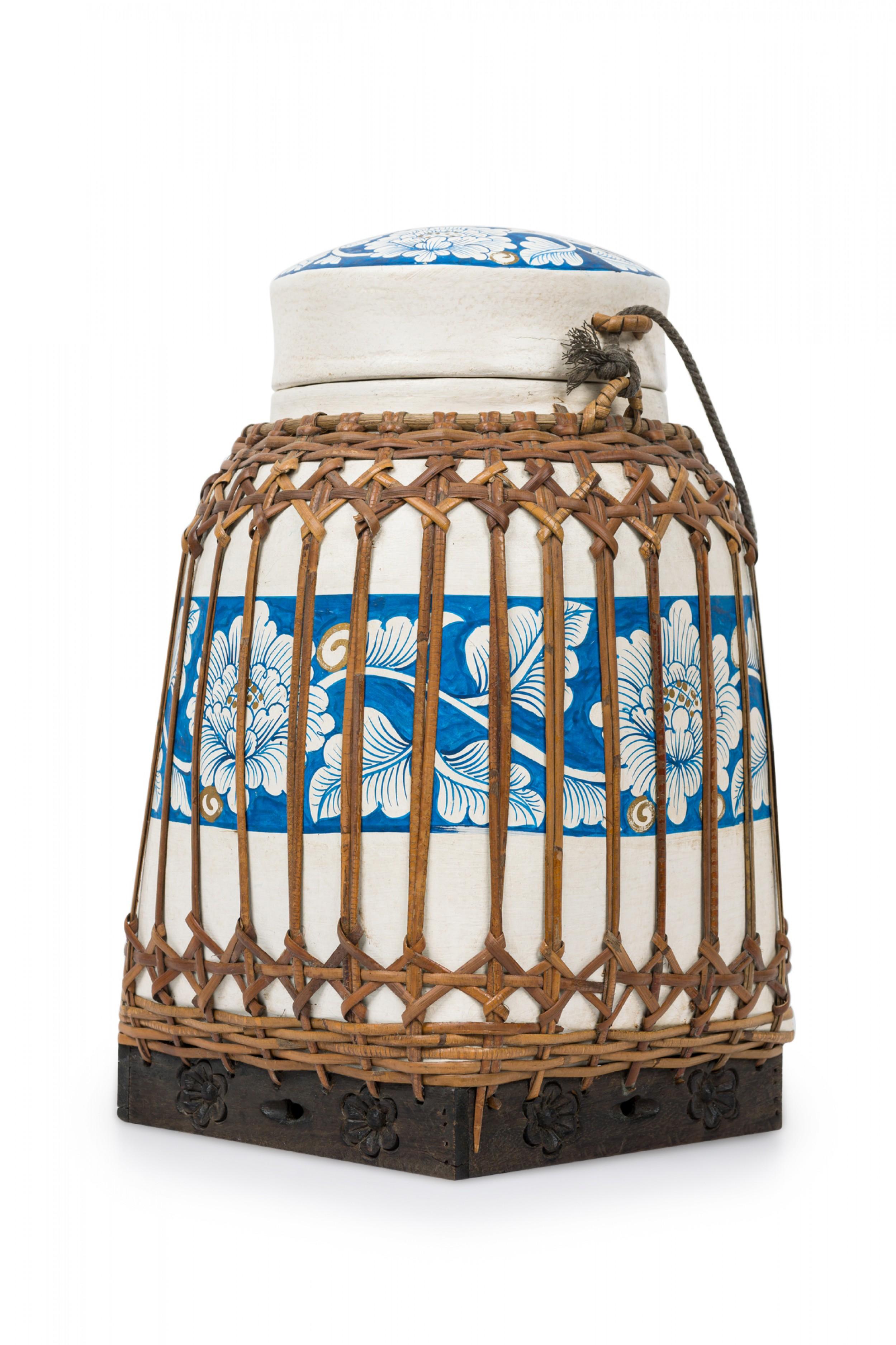 Vintage Chinese papier-mache vessel painted white with a blue and white floral band around the center and top, wrapped in wicker rattan weave, resting on a carved wooden base.